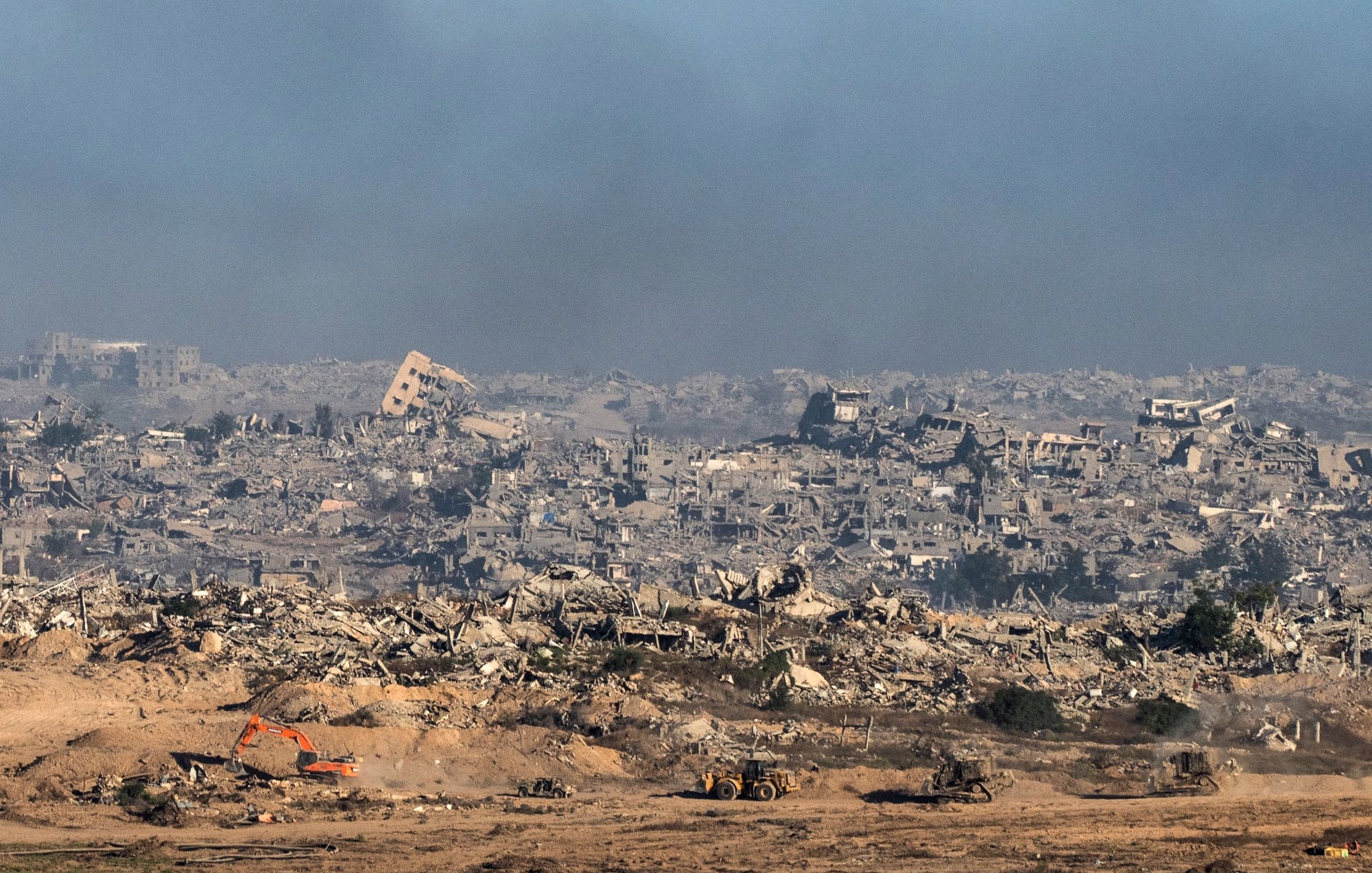 منظره‌ای از ویرانی وسیع، ساختمان‌های تخریب شده و دود در آسمان، با ماشین‌آلات ساختمانی در حال کار.
