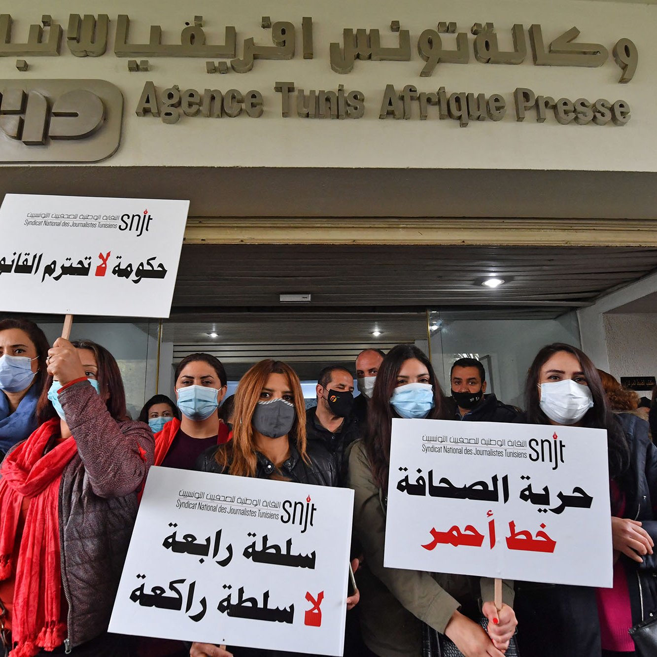 L'image montre un groupe de femmes et d'hommes rassemblés devant un bâtiment, portant des pancartes. Les pancartes expriment des messages liés à la liberté de la presse et critiquent le gouvernement. Les manifestants portent des masques, probablement en raison de préoccupations sanitaires. En arrière-plan, on peut voir le logo d'une agence de presse. L'atmosphère semble déterminée, avec un engagement visible pour des causes liées à la liberté d'expression.