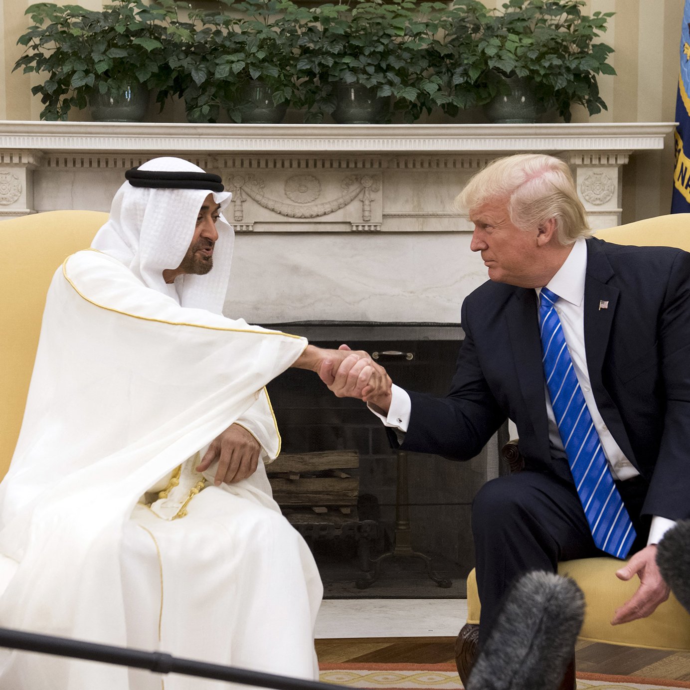 L'image montre deux hommes en train de se serrer la main dans un bureau. L'un porte une tenue traditionnelle, probablement un kandoura et un chapeau, tandis que l'autre est en costume avec une cravate bleue. Ils semblent être en discussion dans un cadre formel, probablement dans un bureau gouvernemental, avec des plantes et des drapeaux visibles en arrière-plan.