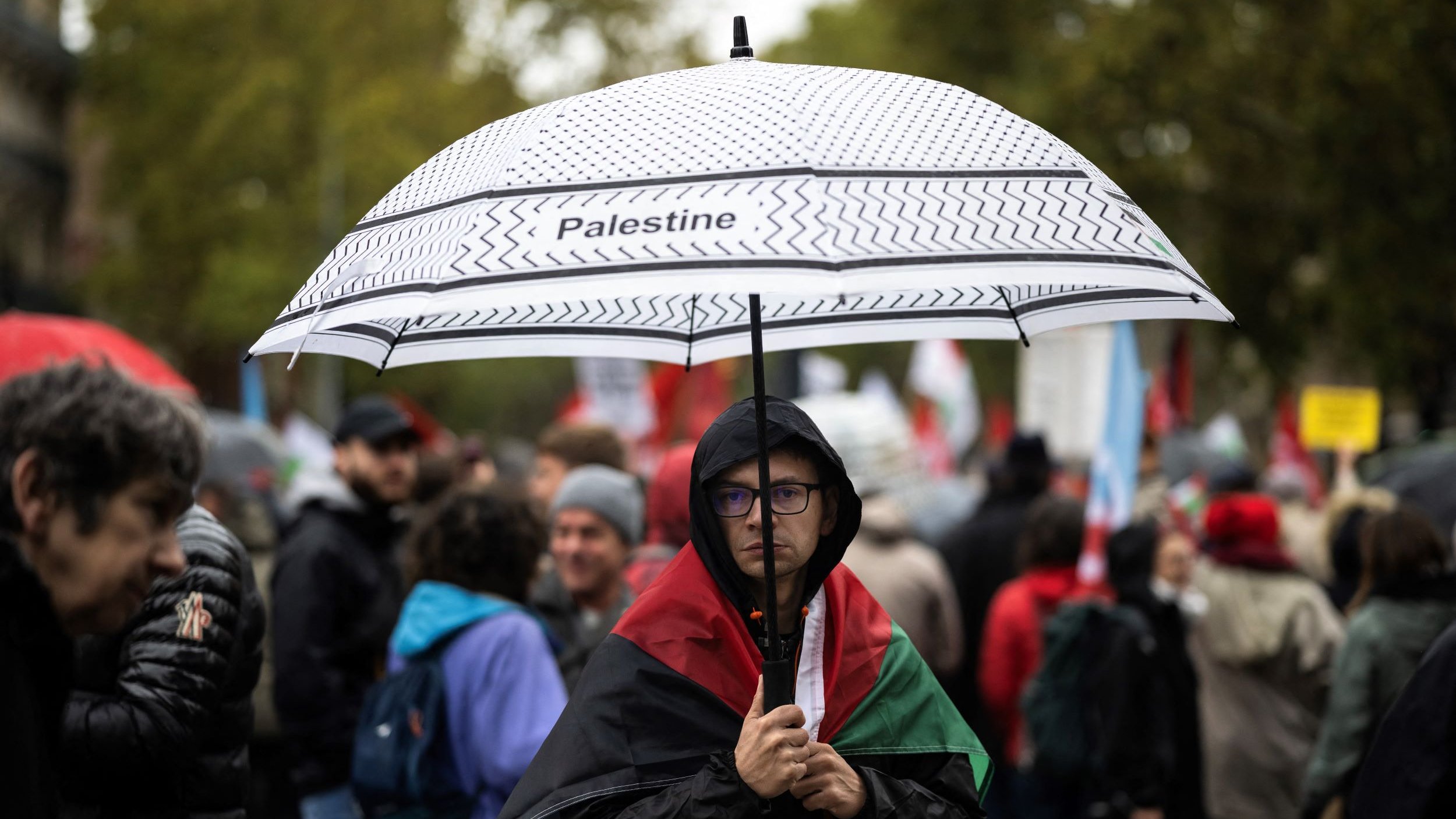 في الصورة، يظهر شخص يحمل مظلة مكتوب عليها "فلسطين"، ويقف وسط حشود من الناس في تظاهرة. يرتدي الشخص علم فلسطين حول كتفيه، وتبدو بعض وجوه المتظاهرين في الخلفية، مع تأثيرات الطقس الممطر، حيث يحمل البعض مظلات أخرى. تعكس الصورة أجواء التظاهر والدعم للقضية الفلسطينية.