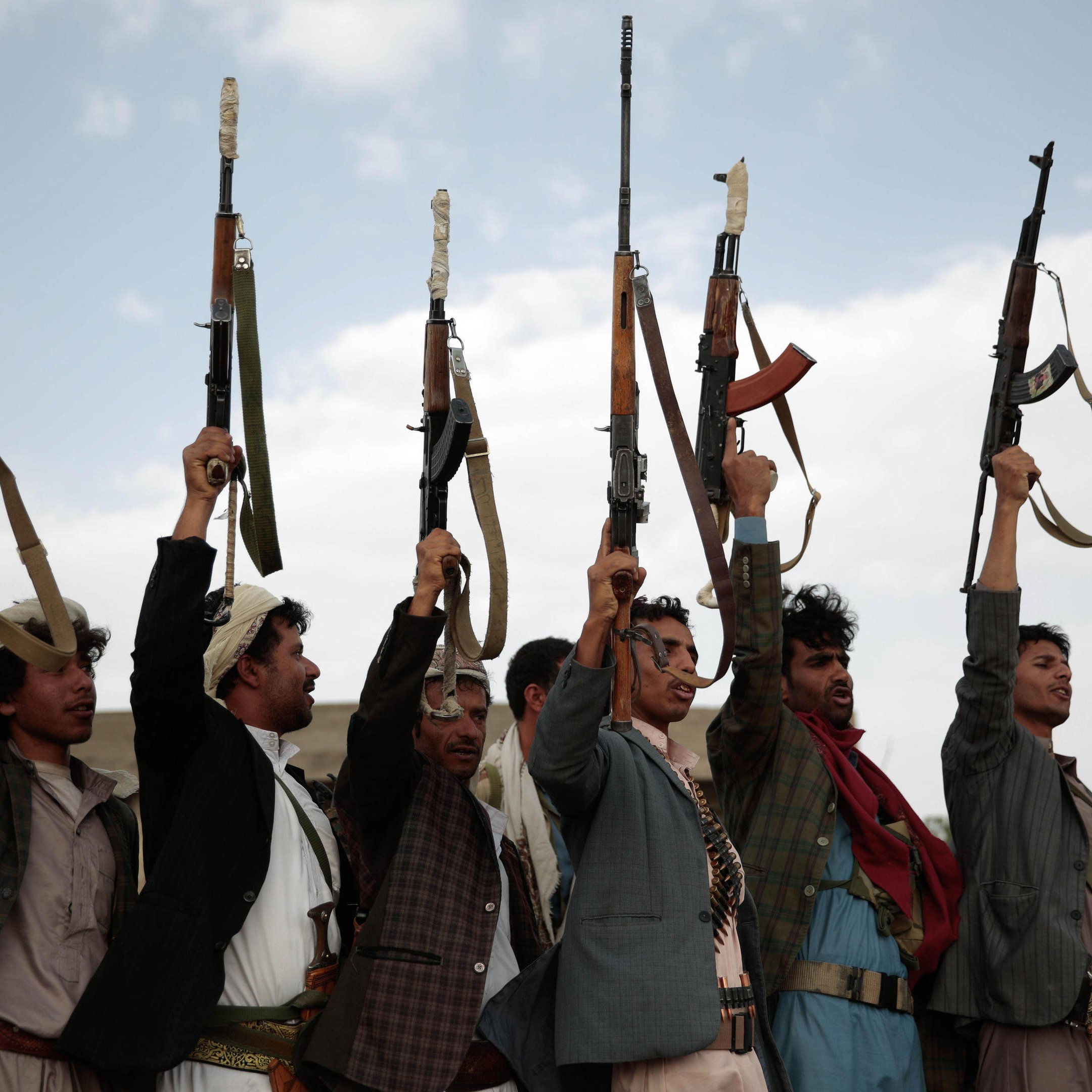 The image shows a group of men holding up rifles in the air. They are standing in a line, with some raising their right arms in a fist while others are focusing on the guns. The setting appears to be outdoors, with a cloudy sky in the background. The men are wearing traditional attire, which varies among them, and there is an overall sense of solidarity and determination in their postures.