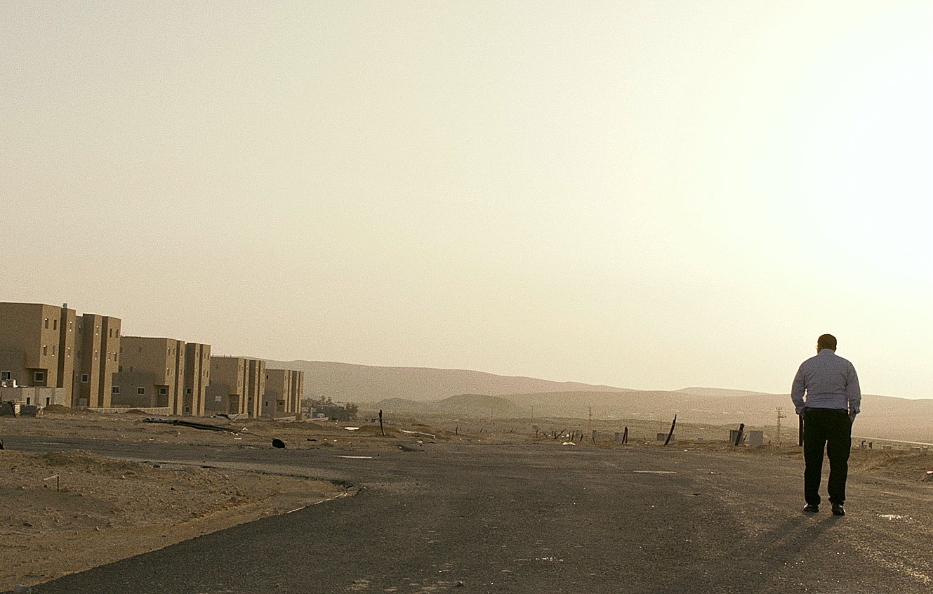 L'image montre un homme marchant seul sur une route déserte. On peut voir en arrière-plan des bâtiments modernes qui semblent inachevés ou abandonnés, avec un paysage désertique s'étendant à l'horizon. La lumière du soleil semble douce, suggérant une ambiance calme et un peu mélancolique. Le ciel est clair, et l'ensemble de la scène évoque une sensation de solitude et d'isolement.