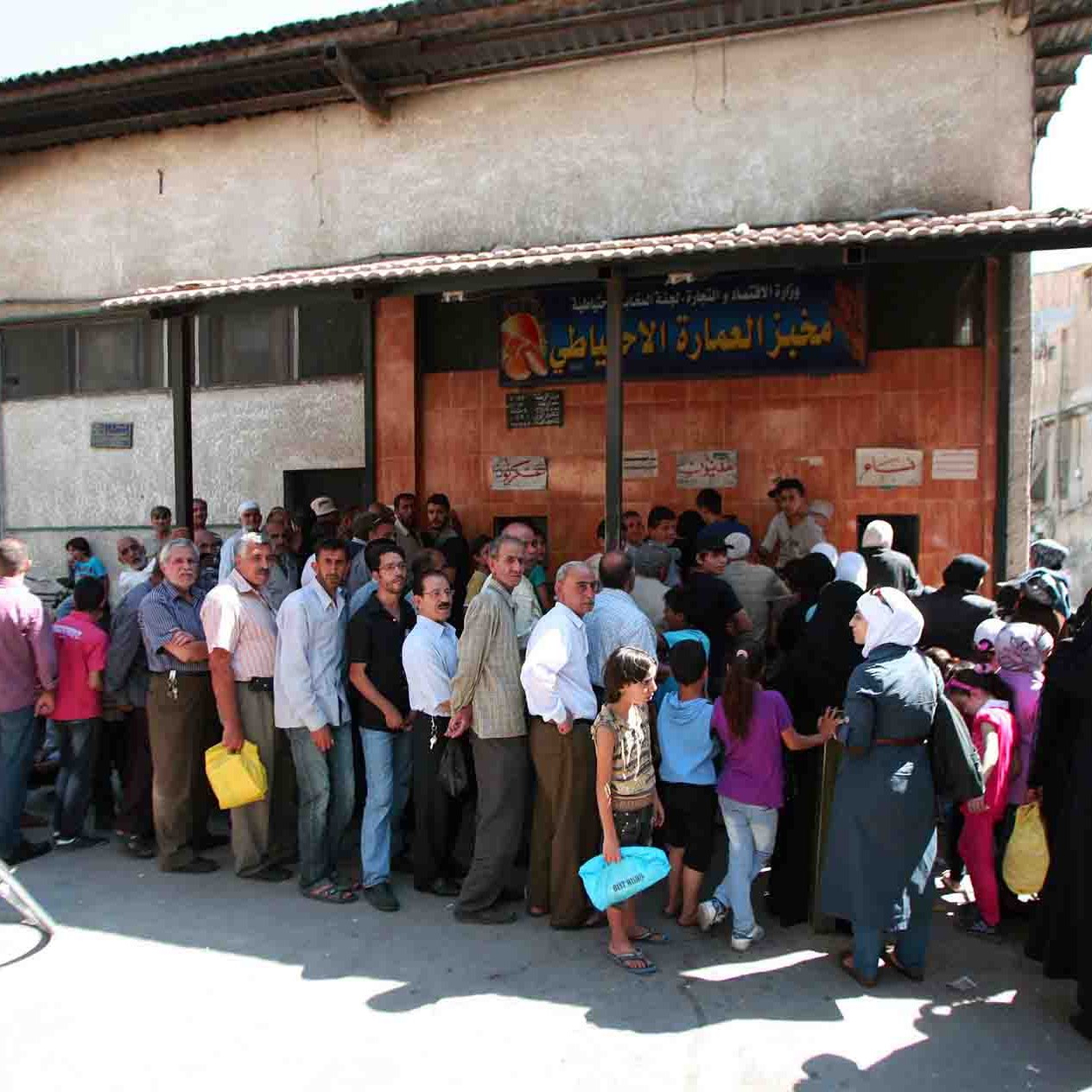 L'image montre une longue file de personnes attendant devant un bâtiment, probablement pour accéder à des services ou des ressources. On peut voir des hommes et des femmes de différentes générations, certains portant des sacs. L'atmosphère semble animée, et le bâtiment est simple, avec une devanture marquée par une enseigne. Des vélos sont stationnés à proximité, et l'environnement urbain est visible en arrière-plan.