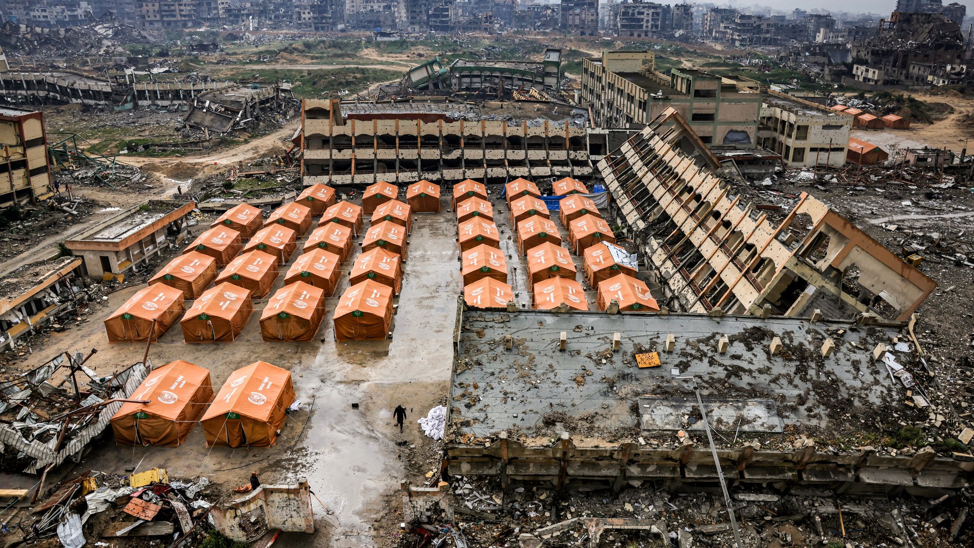 L'immagine mostra un'area devastata, con edifici in rovina sullo sfondo e tende arancioni disposte ordinatamente in primo piano. Il terreno sembra sporco e disordinato, evidenziando una situazione di emergenza o di sfollamento.