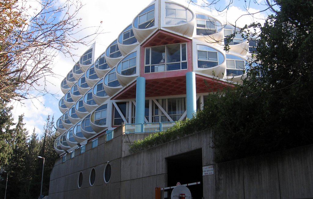 L'image montre un bâtiment moderne avec une architecture unique. Il se compose de plusieurs niveaux, avec une façade caractérisée par des fenêtres en forme de bulle ou de dôme. Les couleurs sont vives, notamment avec des éléments en rouge et en bleu. À l'avant, on peut voir un accès avec un panneau de signalisation et des éléments paysagers comme des arbres sur les côtés. Le bâtiment semble être intégré dans son environnement naturel, avec un design qui attire l'attention.