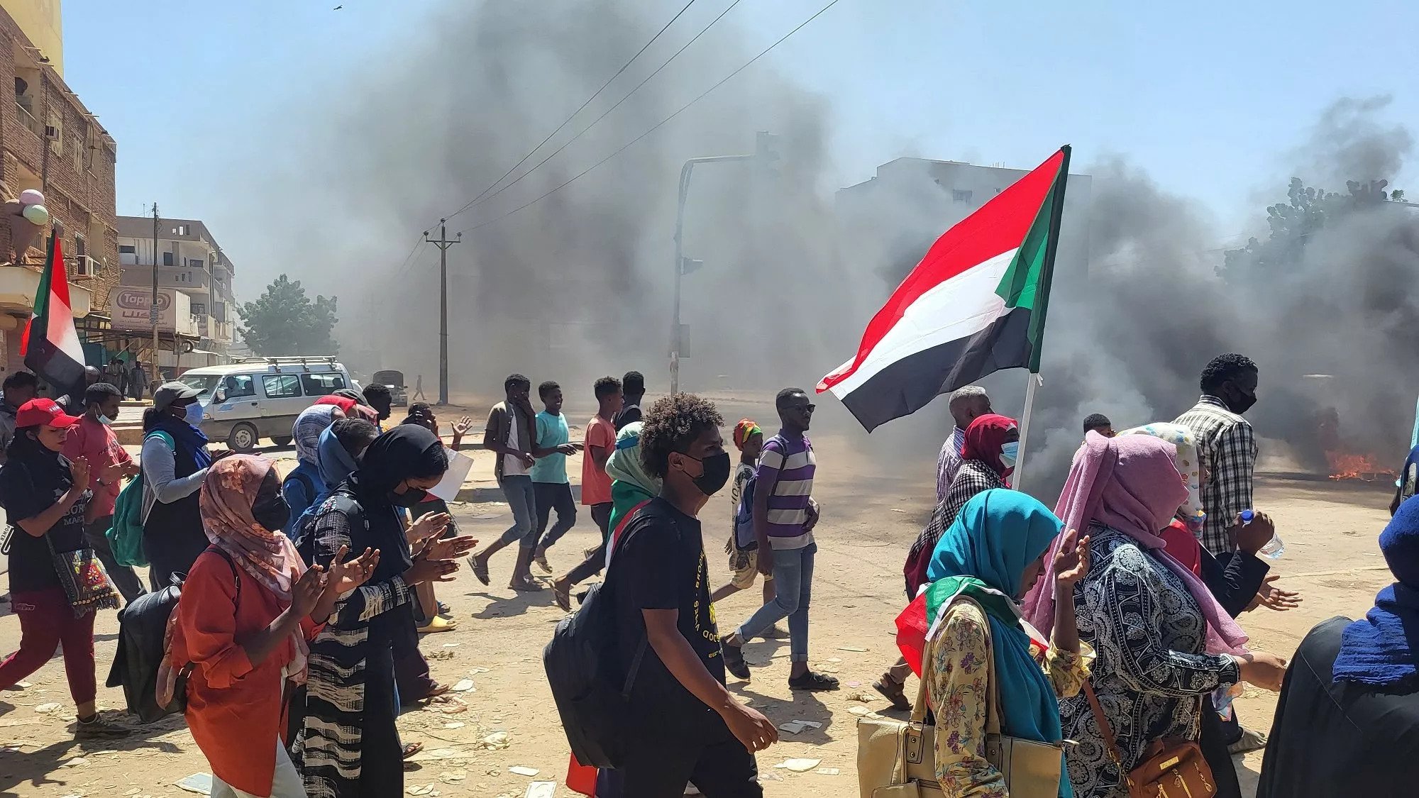 Manifestanti con bandiera del Sudan, in mezzo a fumi di incendi e tensione.