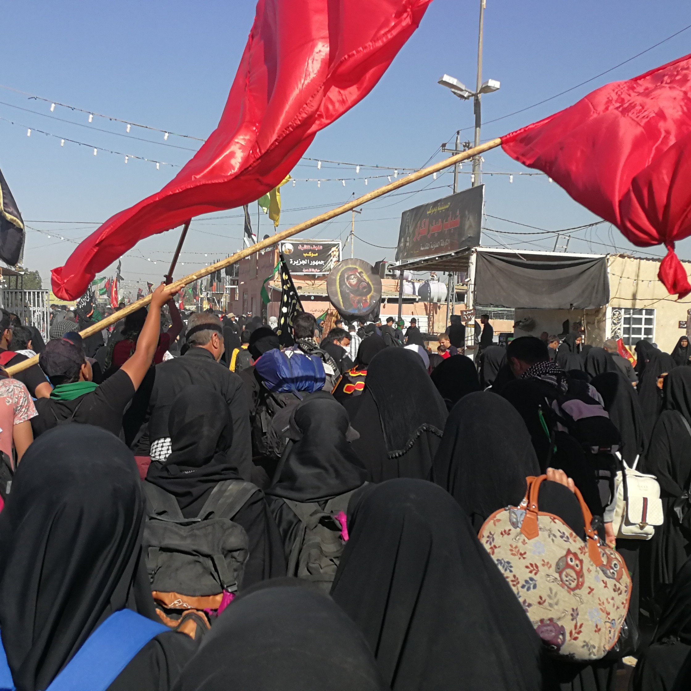 L'image montre une grande foule de personnes rassemblées, principalement vêtues de noir, ce qui suggère un événement commémoratif ou religieux. Plusieurs personnes tiennent des drapeaux rouges, et l'atmosphère semble être celle d'une manifestation ou d'un rassemblement significatif. Les participants portent des vêtements traditionnels et il y a des décors typiques de ce genre d'événement en arrière-plan, comme des bannières et éventuellement des symboles religieux. Le ciel est clair, ce qui indique une journée ensoleillée.