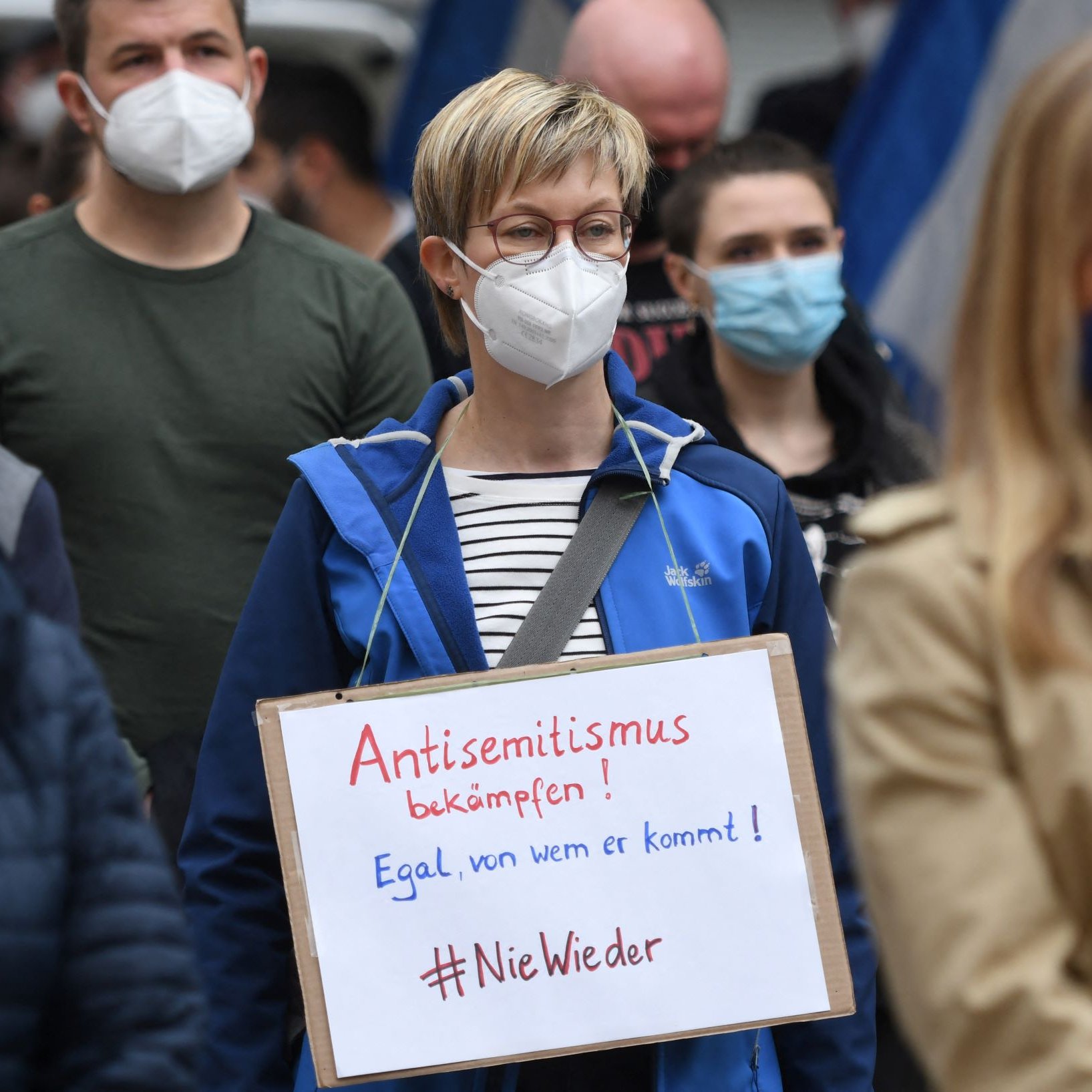 L'image montre une manifestation où des personnes portent des masques. Au premier plan, une femme se distingue avec une pancarte sur laquelle il est écrit en allemand "Antisemitismus bekämpfen! Egal, von wem er kommt! #NieWieder". Elle porte une veste bleue et se tient parmi d'autres manifestants, tous masqués. L'ambiance semble sérieuse et engagée en faveur de la lutte contre l'antisémitisme.