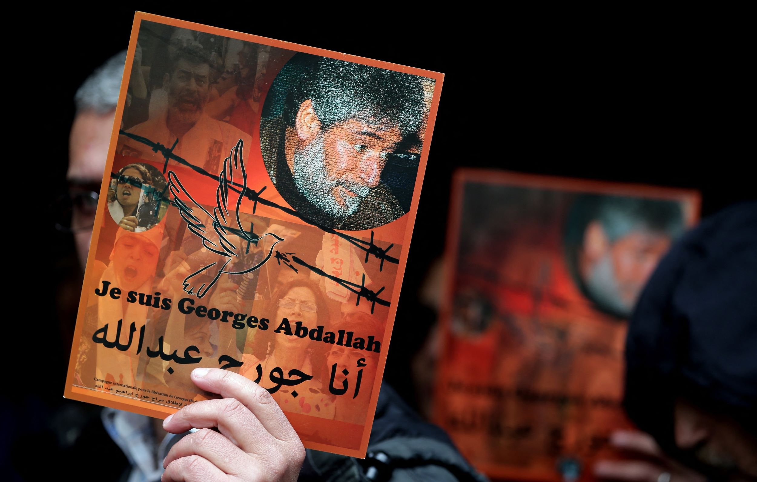 L'image montre une personne tenant une affiche. L'affiche met en avant le message "Je suis Georges Abdallah" en français et en arabe. Elle inclut un portrait de Georges Abdallah entouré de motifs symboliques, comme des barbelés et une colombe. En arrière-plan, on aperçoit des visages et des motifs qui suggèrent un contexte de soutien et d'engagement. L'ambiance semble être celle d'une manifestation ou d'un rassemblement pour la défense de ses droits.