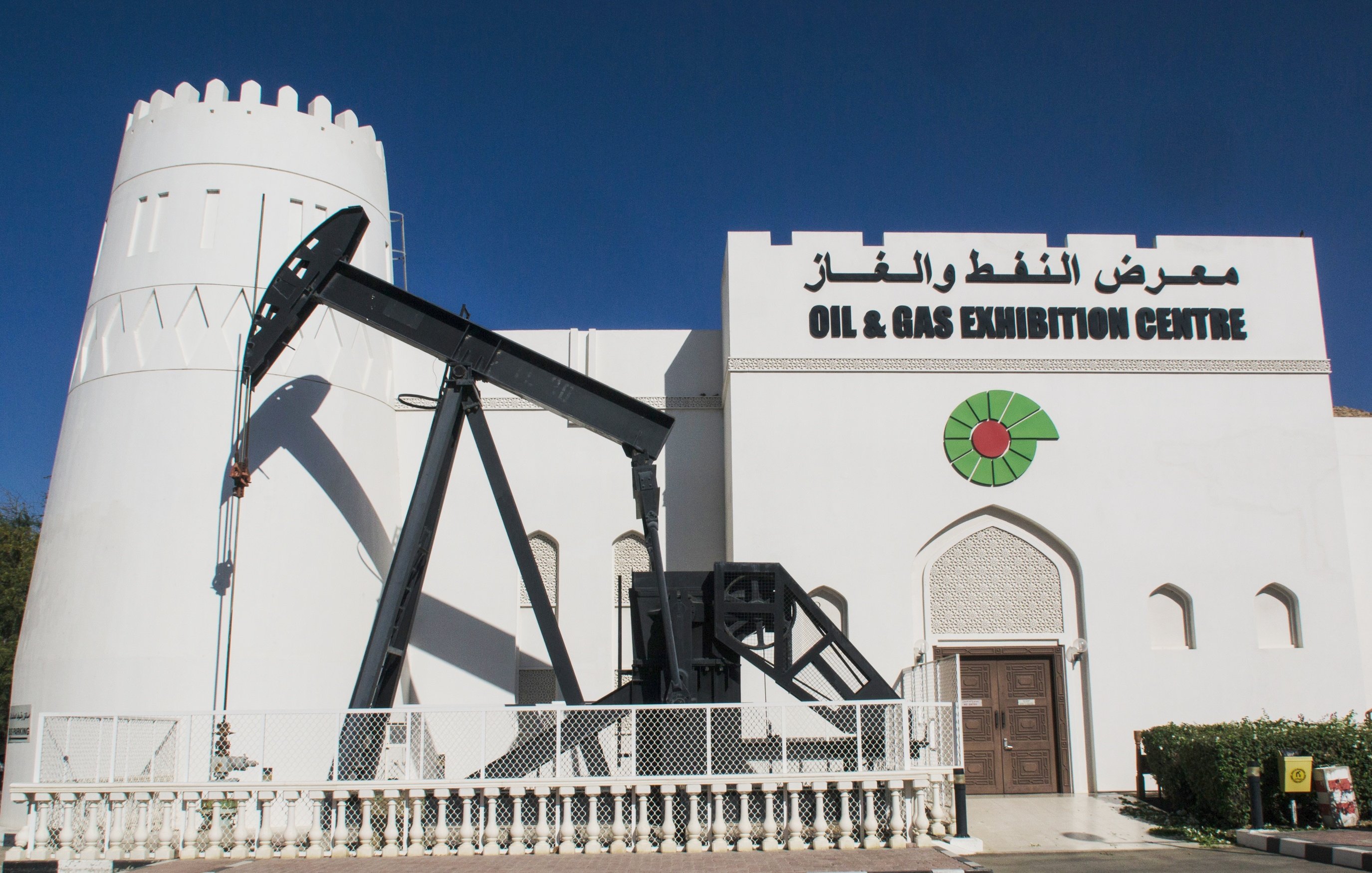 L'image montre un bâtiment blanc qui semble être un centre d'exposition dédié au pétrole et au gaz. À gauche, il y a une structure en forme de pompe à pétrole, typique des installations de forage. Le bâtiment présente un style architectural qui rappelle une forteresse, avec une tour à moitié ronde sur le côté. Au-dessus de l'entrée, on peut voir le nom "OIL & GAS EXHIBITION CENTRE" ainsi que des inscriptions en arabe. La façade est décorée de motifs graphiques, ajoutant une touche esthétique au bâtiment. Le ciel est clair et bleu en arrière-plan.