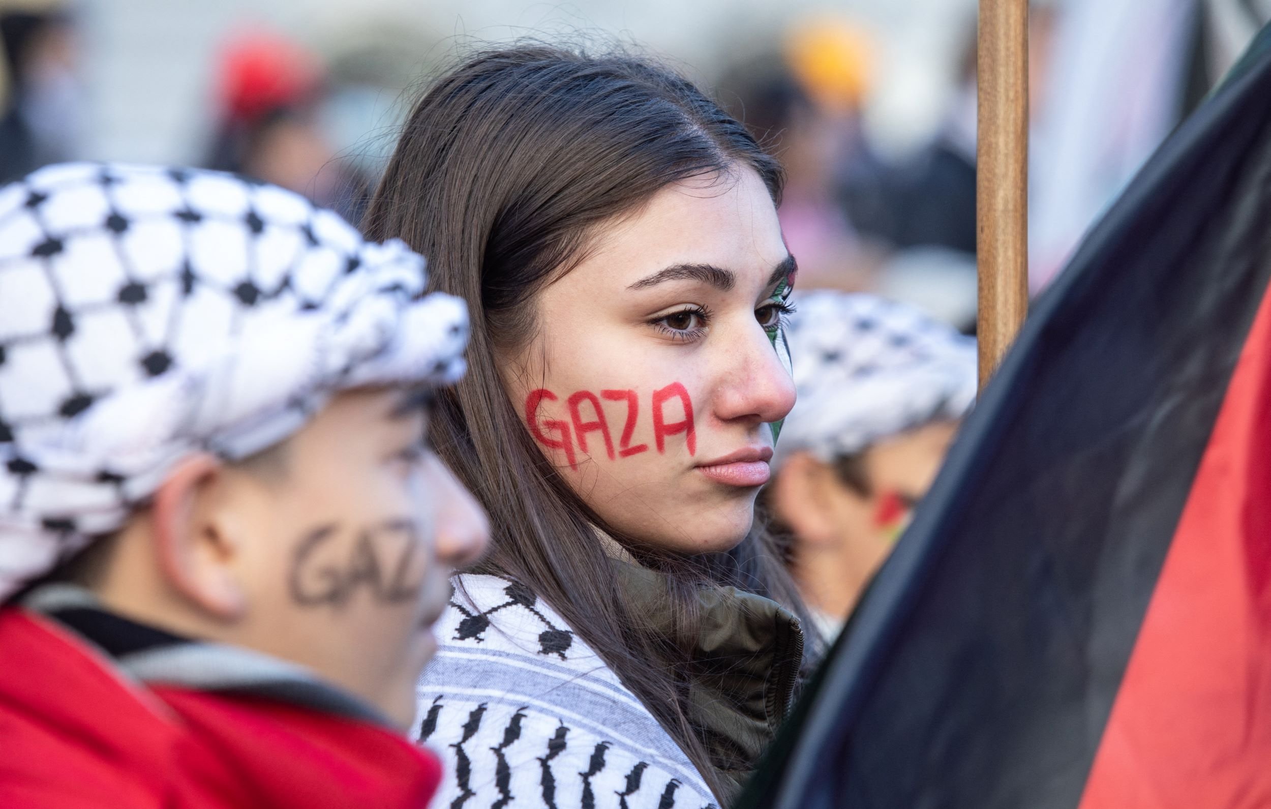 در تصویر، چند نفر در حال شرکت در یک تجمع یا اعتراض به چشم می‌خورند. یک دختر جوان با چهره‌ای جدی و نگران، روی صورتش نوشته‌ای به رنگ قرمز دارد که "Gaza" به نمایش گذاشته شده است. او همچنین شال یا لباسی با طرح‌های خاص به تن دارد. در کنار او، پسرکی نیز دیده می‌شود که روی صورتش نوشته‌ای مشابه به رنگ سیاه دارد. فضای کلی تصویر نشان‌دهنده شور و نشاط در حمایت از مسئله‌ای است که به نظر می‌رسد به غزه مربوط می‌شود. دیگر افراد نیز در پس‌زمینه حضور دارند و فضا به وضوح یک تجمع اجتماعی را تداعی می‌کند.