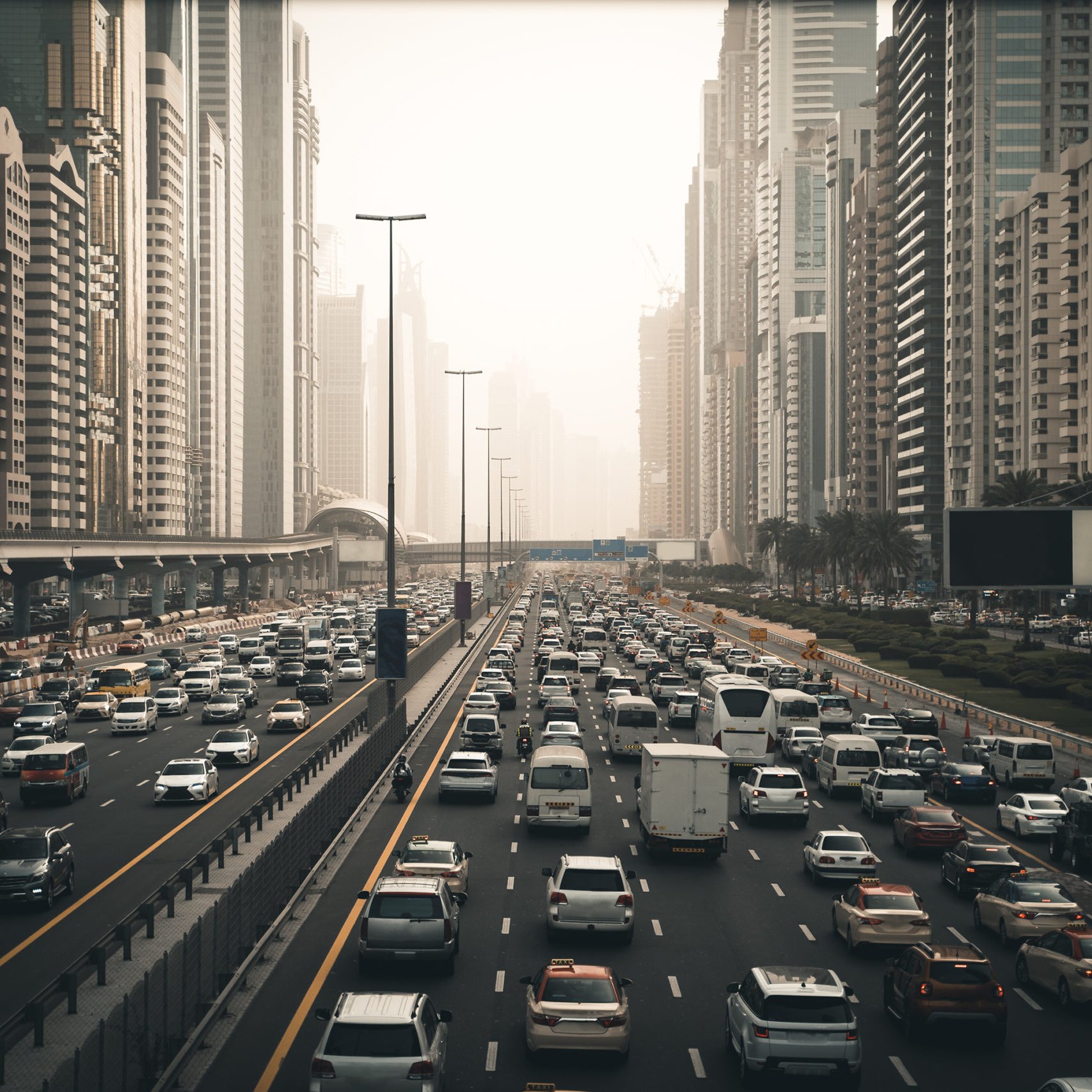 تظهر في الصورة مشهدًا حضريًا لمدينة مزدحمة، حيث تتواجد الكثير من السيارات المتوقفة في زحام مروري على عدة مسارات. تحيط بالمكان ناطحات السحاب العالية التي تدل على التطور العمراني في المنطقة. تبدو السماء مغشاة بضباب خفيف، مما يعطي انطباعًا عن أجواء المدينة في وقت متأخر من اليوم أو خلال الصباح الباكر. هناك مساحات من الأشجار على جوانب الطريق تضفي لمسة من الطبيعة وسط الحضر.