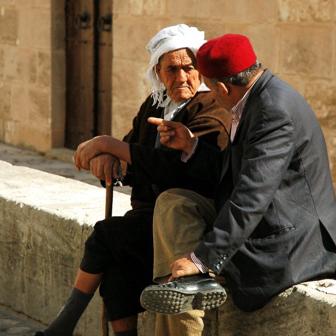Deux hommes âgés discutent assis sur un mur en pierre, l'un portant un fez rouge.