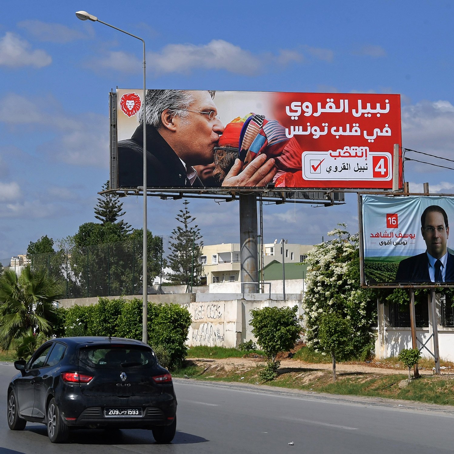 Sur cette image, on voit un panneau publicitaire en arabe. Le panneau montre deux personnages qui s'embrassent, entourés de couleurs vives et de slogans. En bas, on peut voir une photo d'une autre personne (peut-être un candidat) avec un texte supplémentaire à son propos. La scène se déroule dans un environnement urbain, avec des voitures circulant sur une route. Le ciel est bleu, parsemé de nuages.