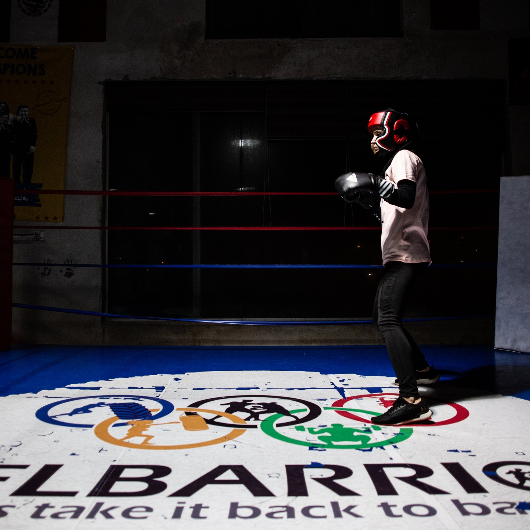 L'immagine mostra un giovane pugile che indossa i guantoni e un casco protettivo, mentre si allena in un ring da boxe. Lo sfondo è parzialmente illuminato, mettendo in risalto il pugile in azione. Sotto di lui, si trova un logo con i simboli olimpici e la scritta "ELBARRIO", che suggerisce un ambiente dedicato allo sport e alla formazione. La scena ha un'atmosfera di concentrazione e determinazione.