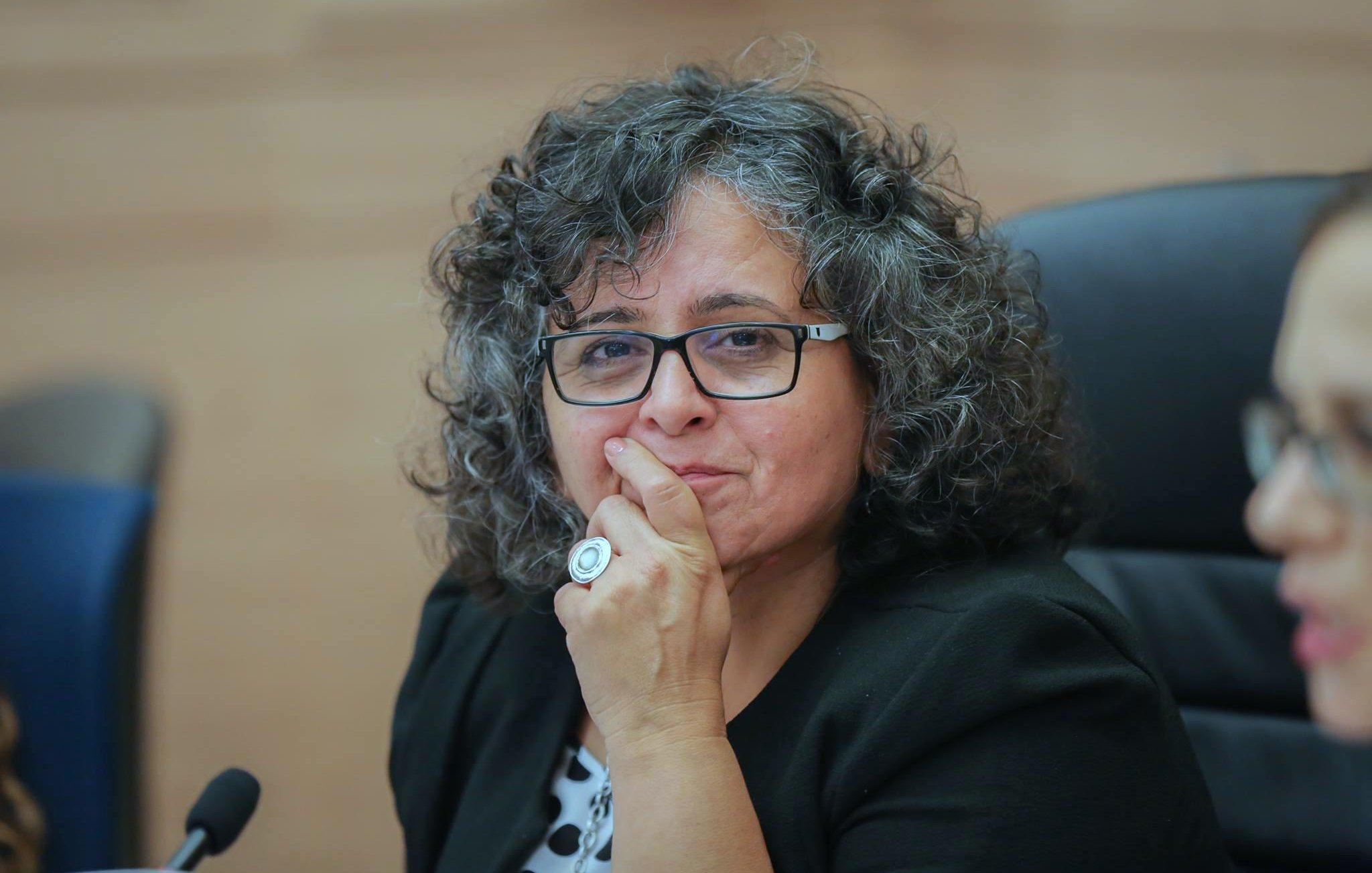 L'image montre une femme avec des cheveux bouclés et un regard réfléchi. Elle porte des lunettes et une veste. Une main est placée sur son menton, ce qui suggère qu'elle est en train de penser ou de considérer une question. L'environnement semble être une salle de réunion ou une assemblée, avec des chaises et une table en arrière-plan.