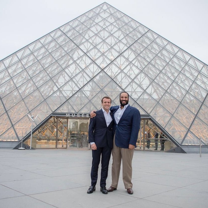 L'image montre deux hommes posant ensemble devant le Musée du Louvre, qui est célèbre pour sa pyramide en verre. Le bâtiment est moderne et impressionnant, contrastant avec l'architecture classique des ailes du musée. Les hommes sourient et portent des costumes formels, se tenant côte à côte, tandis que le ciel est légèrement nuageux, ajoutant une atmosphère calme à la scène.