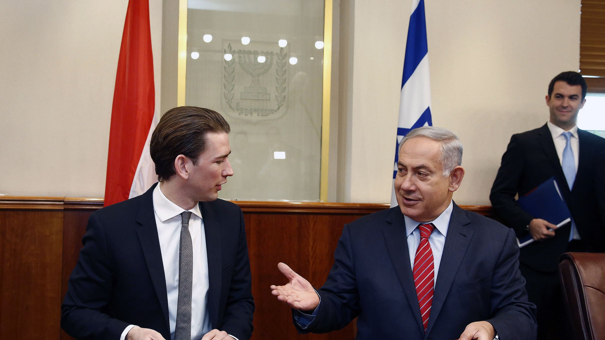 در این تصویر، دو مرد در حال گفتگو و گفت‌وگو هستند. یکی از آنها در سمت چپ، کت و شلوار تیره و کروات دارد و ظاهراً جوانتر به نظر می‌رسد. مرد دیگر در سمت راست، کت و شلوار رسمی به تن دارد و با حرکات دست خود در حال بیان مطلبی است. در پس‌زمینه، پرچم‌هایی به چشم می‌خورند و فضایی رسمی به نظر می‌رسد. نمای کلی تصویر نشان‌دهنده یک جلسه یا توافق رسمی است.