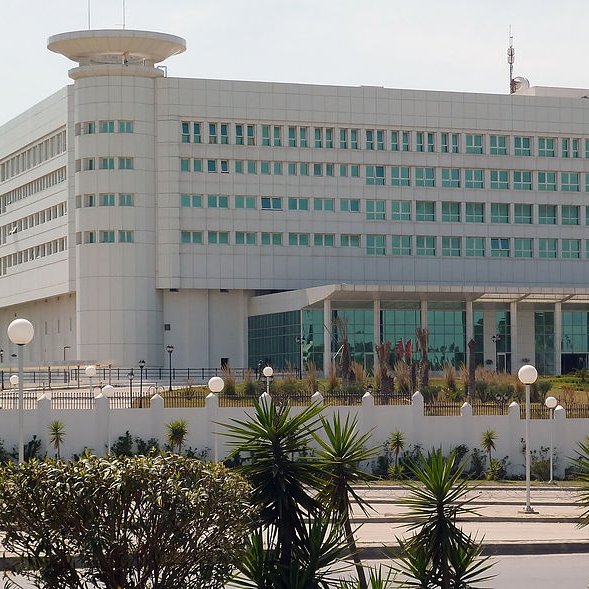 L'image montre un bâtiment moderne et imposant, probablement un centre administratif ou gouvernemental. Il est de couleur blanche avec de grandes fenêtres rectangulaires, donnant une impression de fonctionnalité et de sobriété. Le design architectural est contemporain, avec des formes géométriques claires. À l'avant, il y a un espace ouvert avec des éléments paysagers tels que des plantes et des lampadaires. On peut également voir un drapeau, ce qui pourrait indiquer qu'il s'agit d'un bâtiment officiel. L'environnement semble propre et bien entretenu.