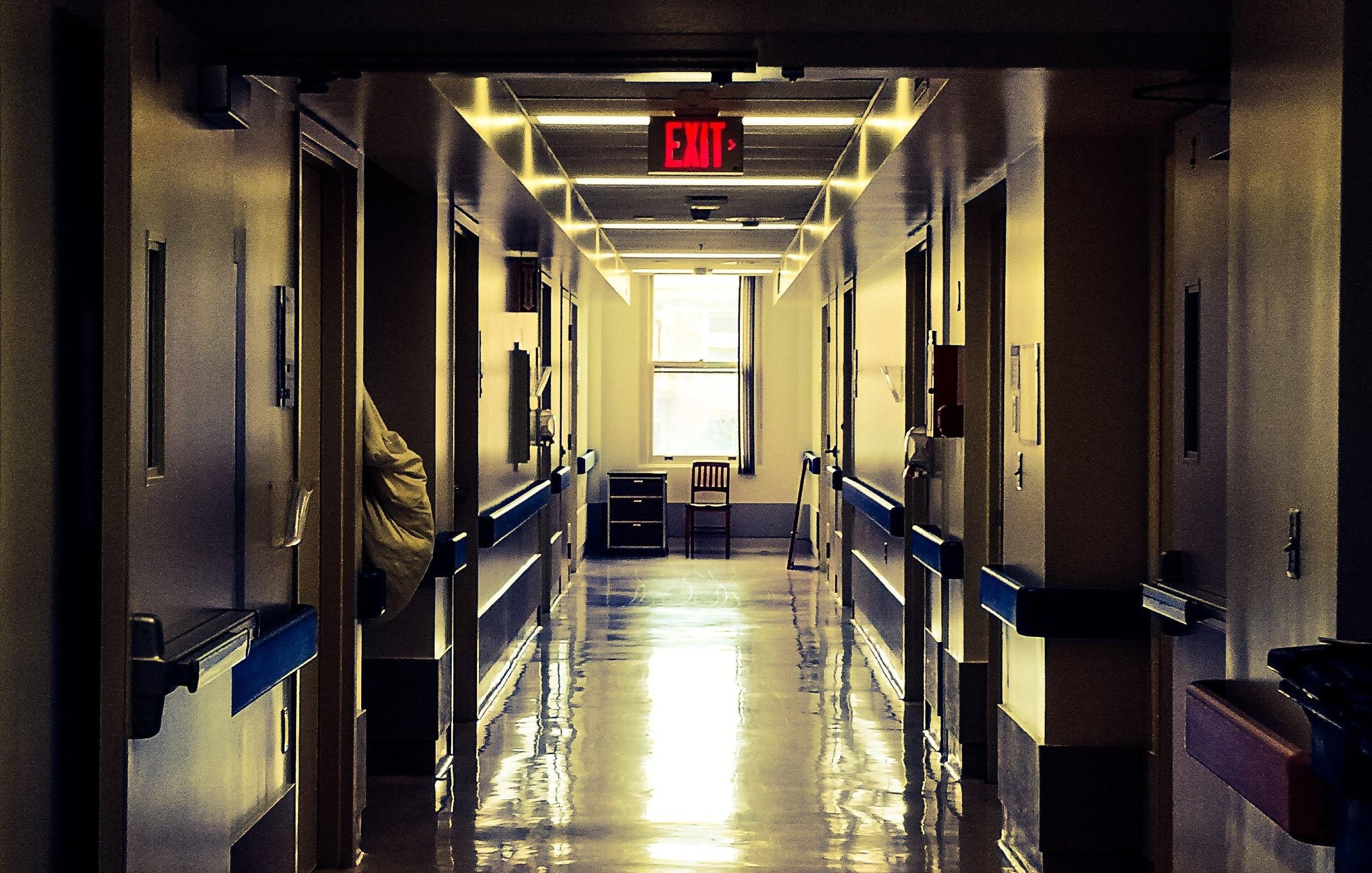 L'image montre un couloir d'hôpital, avec des murs clairs et un sol brillant qui reflète la lumière. Au loin, on aperçoit une fenêtre et une chaise. Les portes sur les côtés sont fermées, et une signalisation de sortie est visible au-dessus. L'ambiance est plutôt calme et légèrement sombre, accentuée par les lumières artificielles.