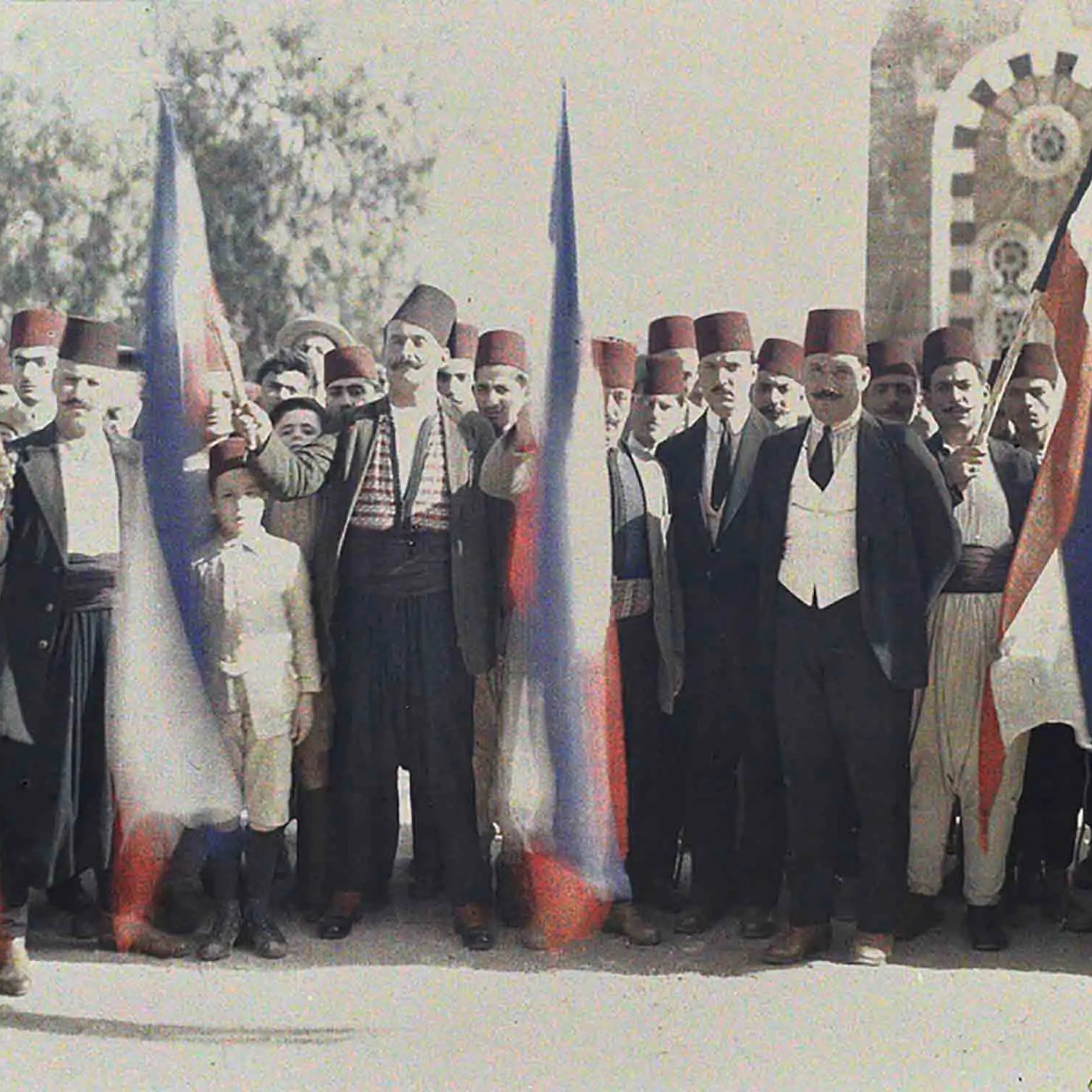 تظهر في الصورة مجموعة من الرجال واقفين في صف واحد، يحملون أعلامًا. يرتدي معظمهم الملابس التقليدية، بما في ذلك الأزياء التي تضم الطربوش الفاسي. يظهر في الصورة أيضًا صبي صغير في وسط المجموعة. يبدو أنهم يحتفلون أو يشاركون في فعالية معينة، حيث يُظهر تعبيرهم الحماس والجدية. الخلفية تحتوي على مبنى قديم، مما يعطي انطباعًا تاريخيًا ومناسبًا للسياق.