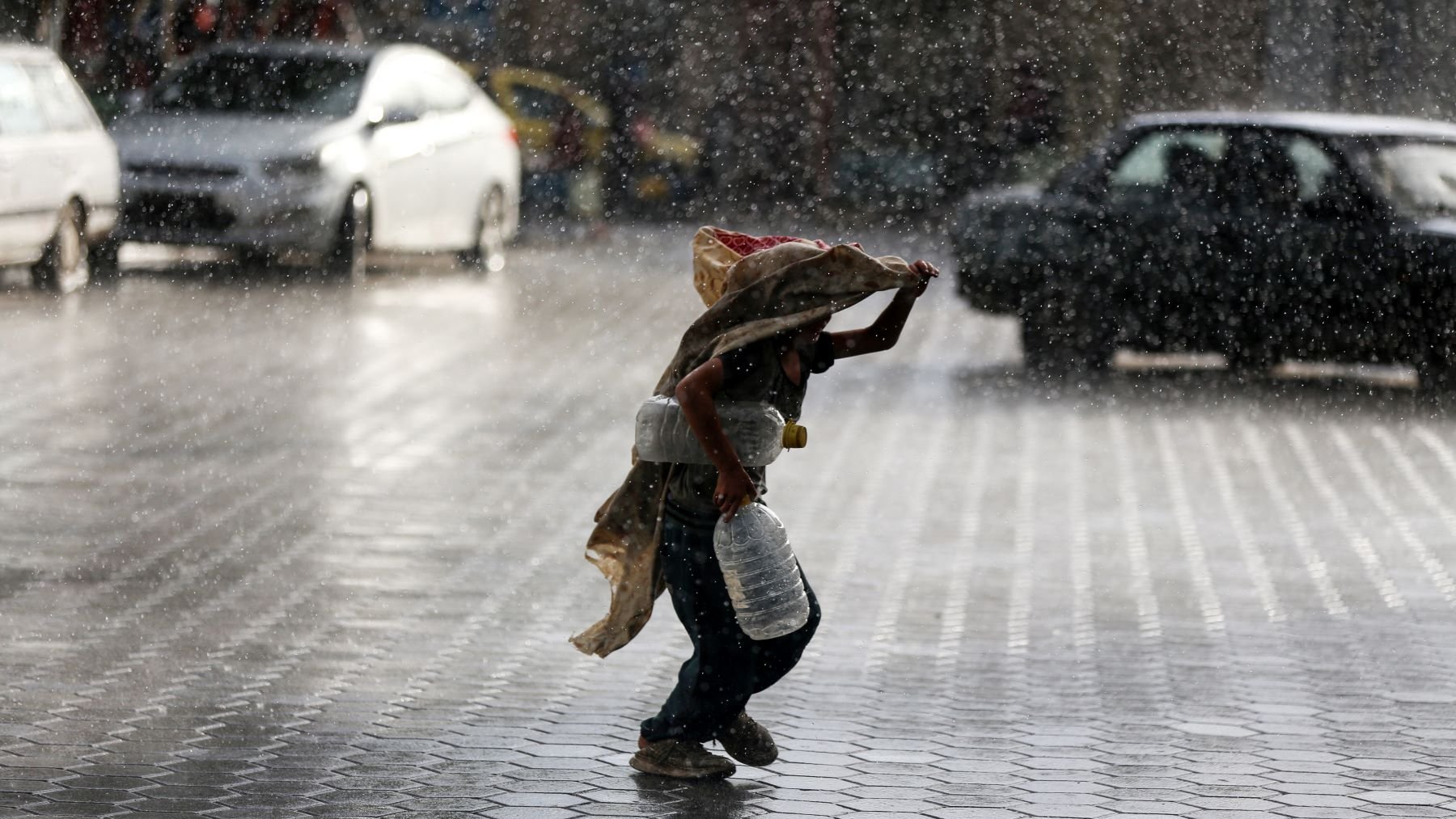 L'image montre un enfant courant sous la pluie intense, portant une écuelle en métal et un sac. Il semble se protéger de la pluie avec un tissu qui couvre sa tête et ses épaules. Le sol est mouillé et réfléchit la lumière, tandis que l'environnement autour de lui est flou en raison des cordes de pluie. Des voitures sont visibles en arrière-plan, accentuant l'atmosphère urbaine de la scène.