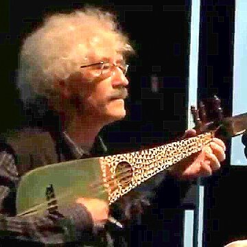 L'image montre un homme âgé avec des cheveux bouclés et gris, jouant d'un instrument de musique à cordes. Il porte des lunettes et une chemise à motifs, ainsi qu'un gilet. On peut voir une ambiance de concert ou de performance, avec un éclairage qui met en valeur l'artiste et son instrument, qui ressemble à un luth.