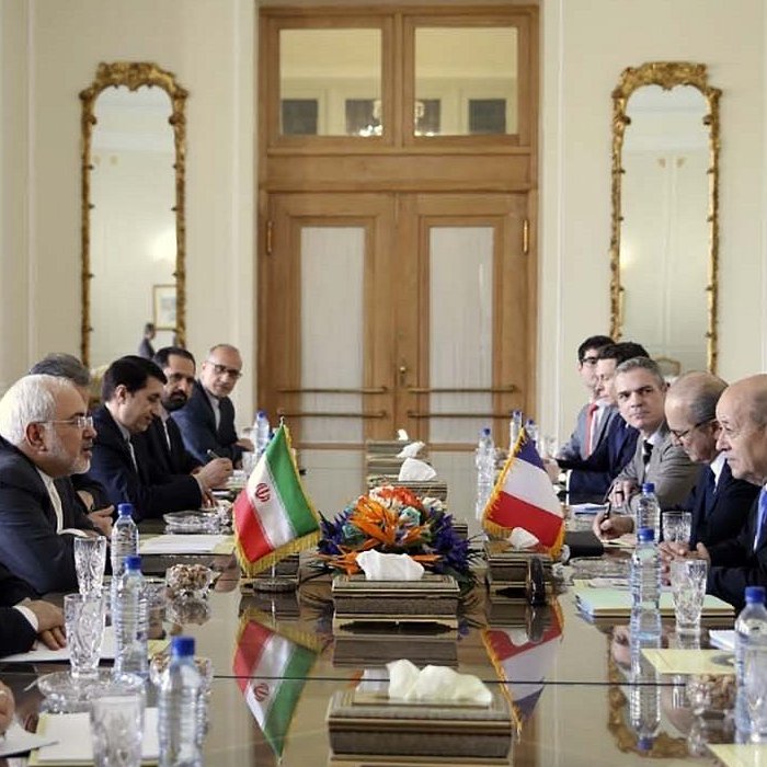 در این تصویر، یک جلسه رسمی در حال برگزاری است. تعدادی از افراد در دو طرف یک میز بزرگ نشسته‌اند و در مرکز میز، پرچم‌های ایران و یک کشور دیگر قرار دارد. برخی از افراد به نظر می‌رسد که مقامات سیاسی یا دیپلمات هستند. فضا شبیه به یک سالن کنفرانس است، با نور طبیعی که از پنجره‌ها وارد می‌شود و دکوراسیون کلاسیک شامل آینه‌ها و تابلوهای هنری. عده‌ای از افراد با دقت به سخنران گوش می‌دهند، در حالی‌که برخی دیگر مشغول نوشتن یا یادداشت‌برداری هستند.