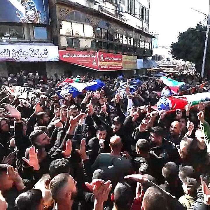 L'image montre une grande foule rassemblée dans une rue, avec des personnes portant des drapeaux et des pancartes. Il y a des cercueils sur des supports, symbolisant un hommage ou des funérailles. La foule est visiblement très engagée, levant les mains et exprimant des émotions fortes. L'environnement urbain est marqué par des bâtiments et des affiches en arrière-plan. L'atmosphère est chargée de gravité et de solidarité.