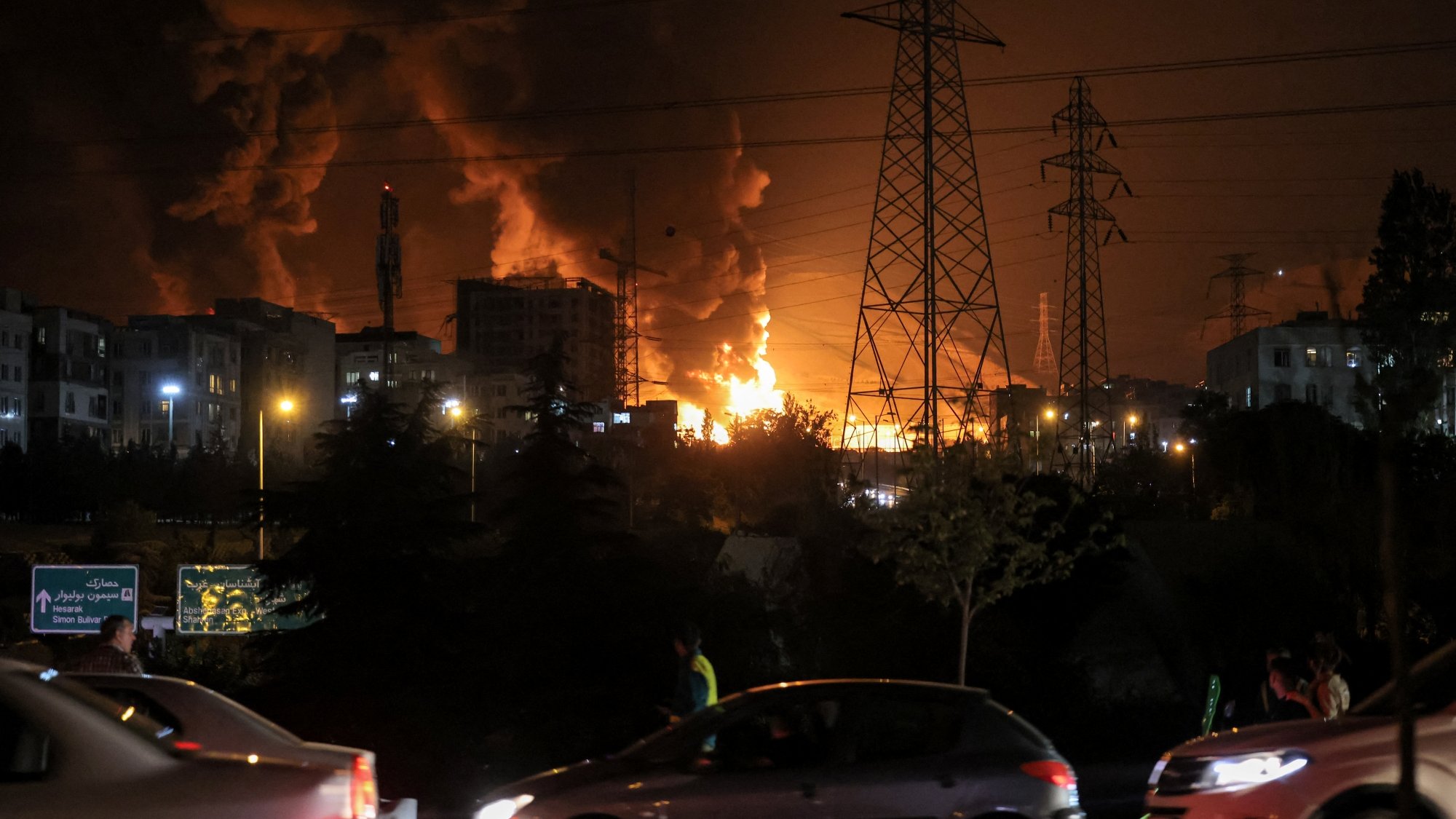 Bright flames and dark smoke rise from buildings at night, with cars passing nearby.