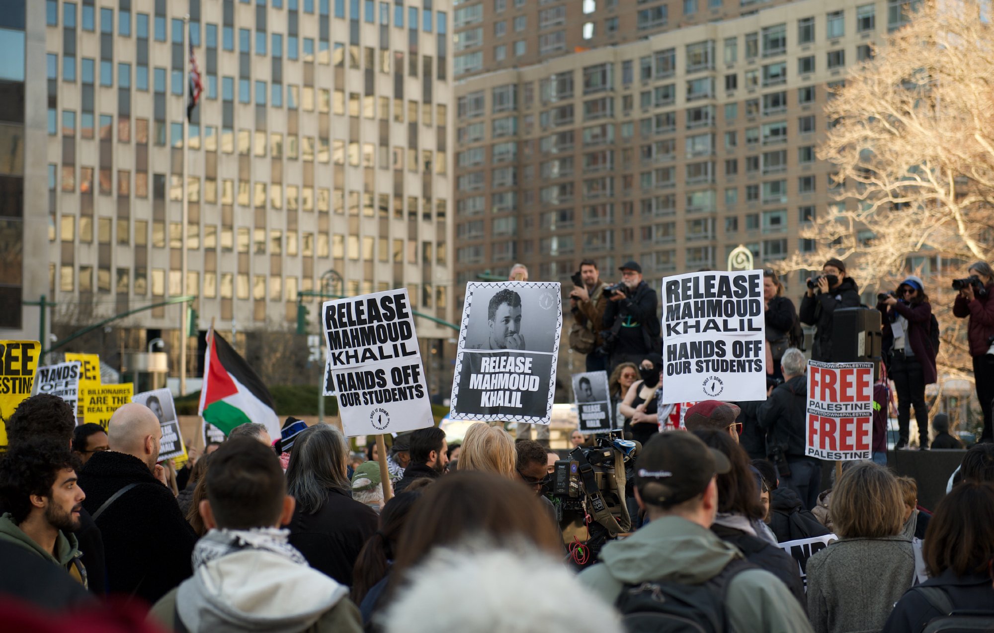 Des manifestants se rassemblent dans une ville, tenant des panneaux avec des slogans, notamment pour la libération de Mahmoud Khalil et des revendications pour les droits des étudiants. En arrière-plan, on peut voir des bâtiments et des personnes utilisant des caméras. L'ambiance est déterminée et engagée.