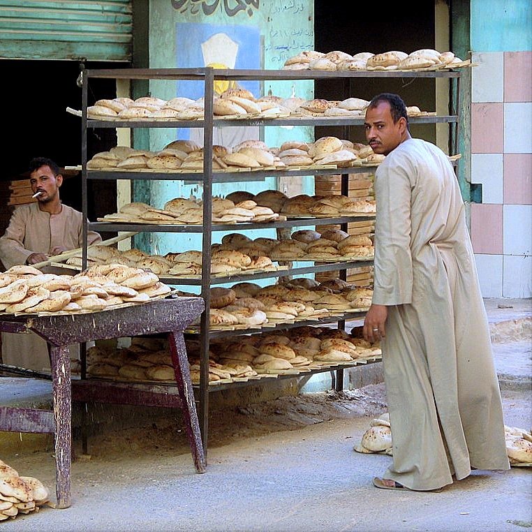 L'image montre une scène de la vie quotidienne dans une boulangerie. Il y a deux hommes : l'un est debout derrière un chariot portant des pains, tandis que l'autre semble en train d'observer ou d'interagir avec des pains empilés sur des étagères. Les pains sont de forme ronde et semblent frais, sortant tout juste du four. En arrière-plan, on aperçoit un mur avec des écritures et des graffiti, donnant une atmosphère typique d'un marché local. Le paysage autour est urbain, avec des bâtiments qui semblent usés par le temps.