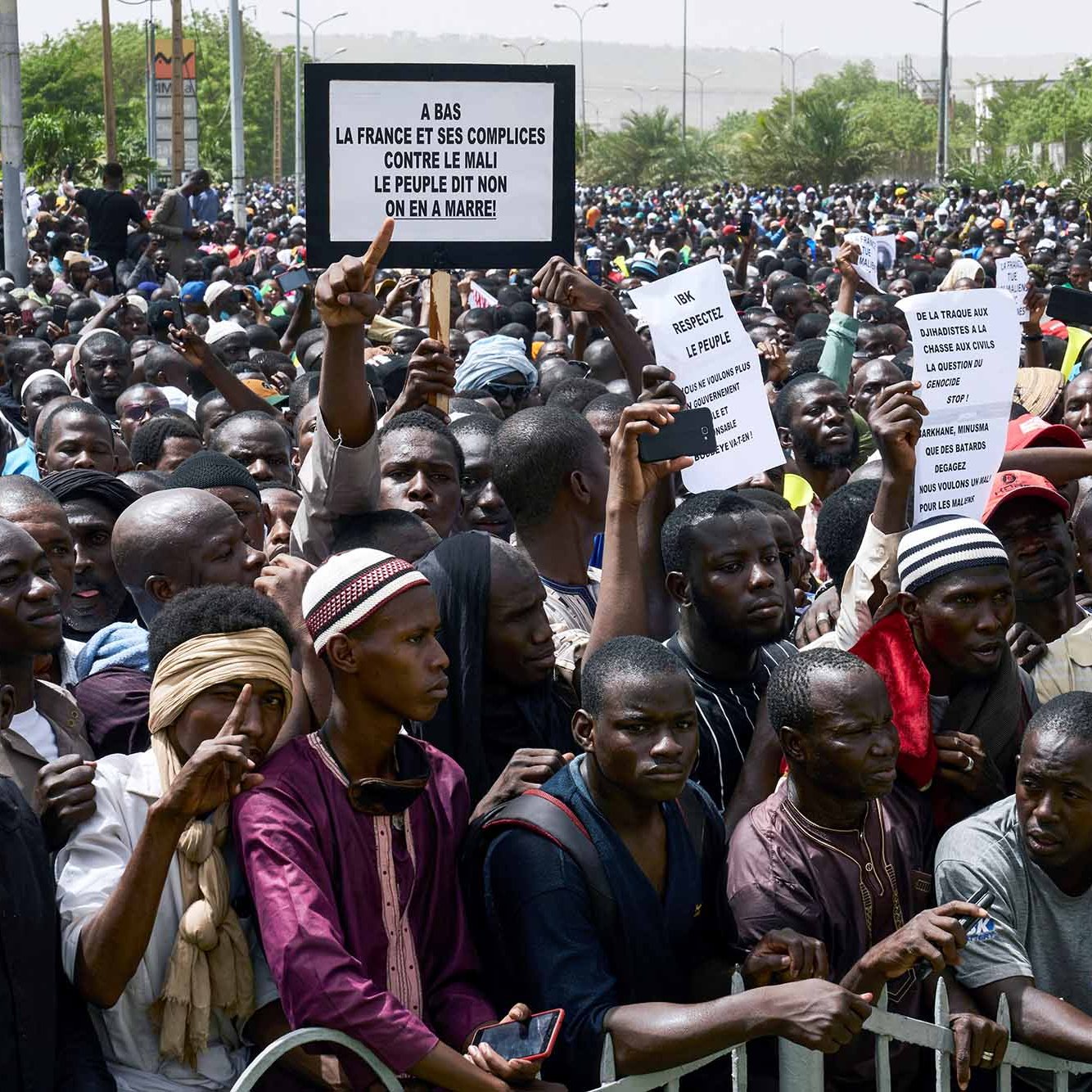L'image montre une grande foule de manifestants réunis, tenant des pancartes. Les personnes semblent exprimer leur mécontentement et leurs revendications. On peut voir une diversité de vêtements parmi les participants, allant des habits traditionnels à des vêtements plus modernes. L'atmosphère paraît énergique et engagée, avec des individus qui tentent de faire entendre leur voix. Des barrières sont présentes pour délimiter l'espace.