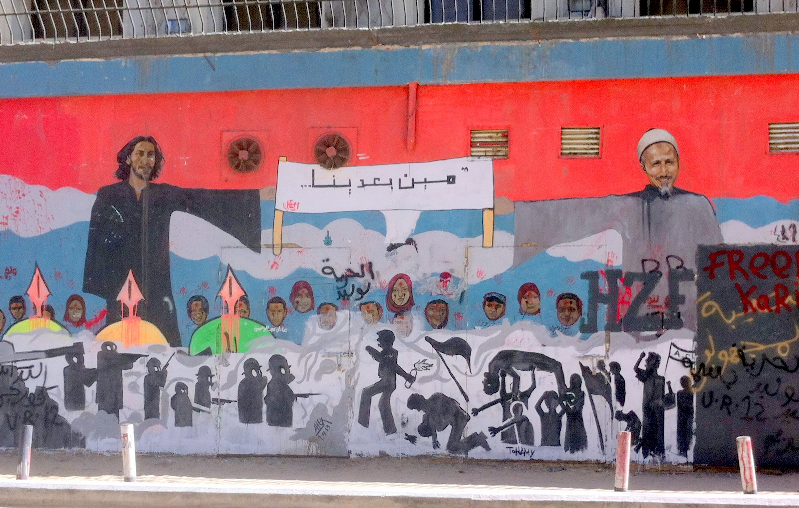 L'image montre une grande fresque murale qui illustre des figures humaines et des symboles de résistance. À gauche, on voit une personne avec de longs cheveux, et à droite, un homme âgé portant un turban. Entre eux, il y a une inscription dans une langue arabe. Le mural est riche en détails, avec des silhouettes de personnes et des flèches, suggérant des thèmes de lutte et d'espoir. Le fond est principalement rouge, créant un contraste fort avec les personnages et les éléments représentés.