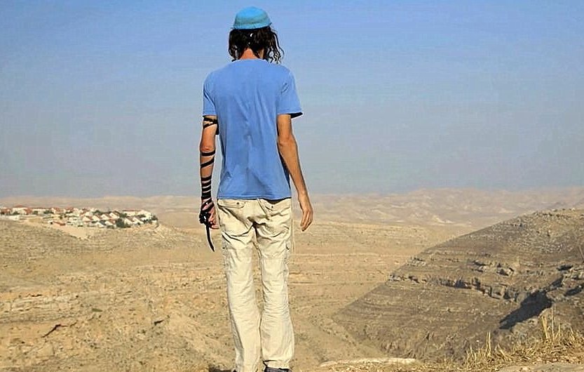 Cette image montre une personne de dos, se tenant au bord d'une falaise. Elle porte un t-shirt bleu et des pantalons clairs, ainsi qu'une kippa sur la tête. Son bras gauche est vêtu d'un bandage. Le paysage environnant est désertique, avec des collines et une vallée visible à l'arrière-plan. On peut apercevoir quelques maisons au loin, suggérant une présence humaine dans cette vaste étendue. L'atmosphère est calme, avec un ciel clair.