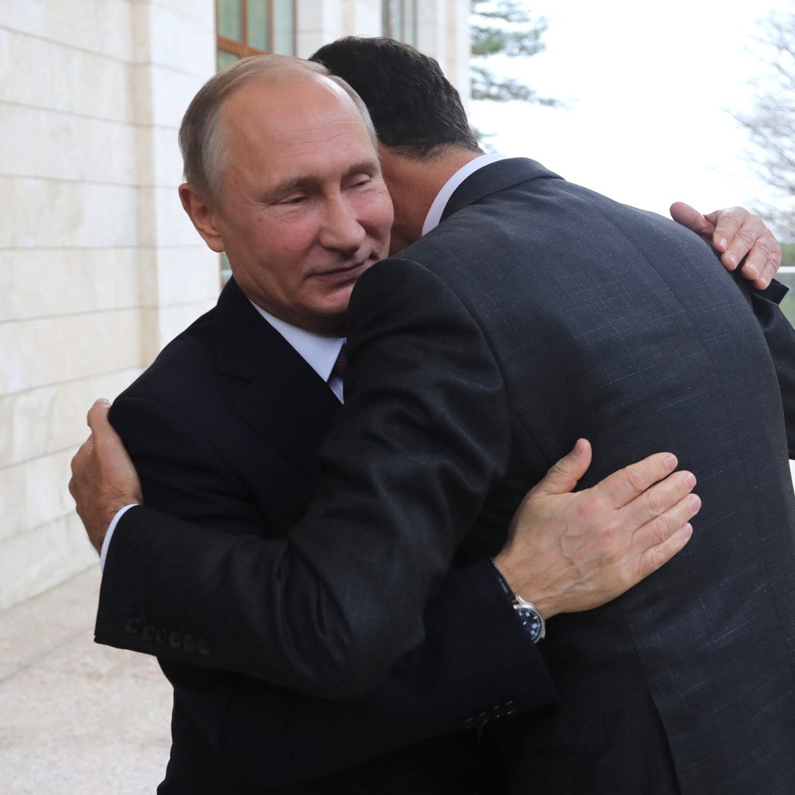 L'image montre deux hommes s'enlaçant dans un cadre officiel. L'un d'eux porte un costume foncé et semble sourire légèrement, tandis que l'autre, également en costume, se penche pour l'étreindre. L'environnement semble être d'une architecture moderne, avec des murs en pierre et un éclairage naturel. L'atmosphère paraît chaleureuse malgré le contexte formel.