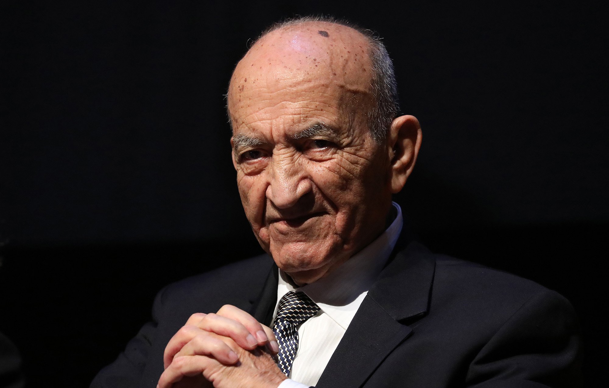 L'image montre un homme âgé, portant un costume noir et une cravate rayée. Il a une expression sérieuse sur son visage, avec un léger sourire. Ses mains sont posées l'une sur l'autre devant lui, et il semble être en train de réfléchir ou d'écouter attentivement. Le fond est sombre, ce qui met en valeur son visage et sa tenue.