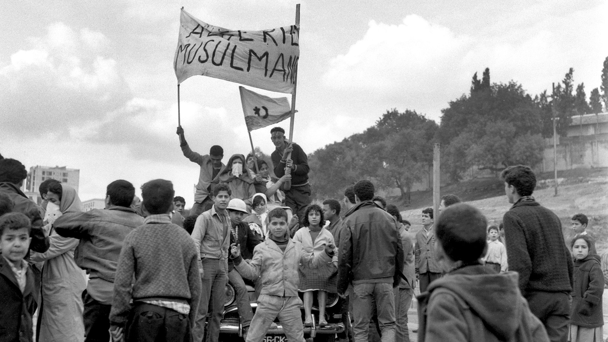 L'image montre une scène de rue animée, probablement lors d'une manifestation ou d'un rassemblement. Des groupes de personnes, en majorité des jeunes, se rassemblent autour d'une voiture. Certains tiennent des drapeaux ou des banderoles sur lesquelles des messages sont affichés, exprimant des revendications. L'atmosphère semble dynamique et chargée d'émotion, avec des visages montrant diverses réactions. En arrière-plan, on aperçoit un paysage urbain avec des arbres et des bâtiments. Le noir et blanc de la photo accentue le caractère historique et sérieux de la scène.