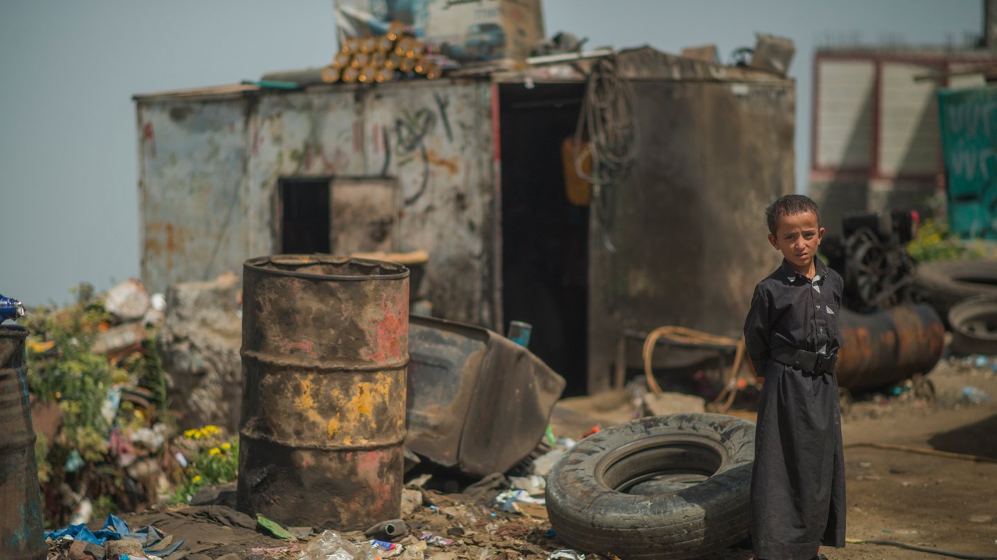 L'image montre un enfant debout dans un environnement désolé. Il porte des vêtements sombres, semblant se tenir près d'une zone remplie de débris, y compris des pneus usés et des barils. En arrière-plan, on aperçoit une structure rustique qui semble être une sorte d'abri. Le sol est sale, jonché de déchets, reflétant une atmosphère de pauvreté et de négligence. L'enfant a une expression sérieuse, ce qui souligne le caractère difficile de son environnement.