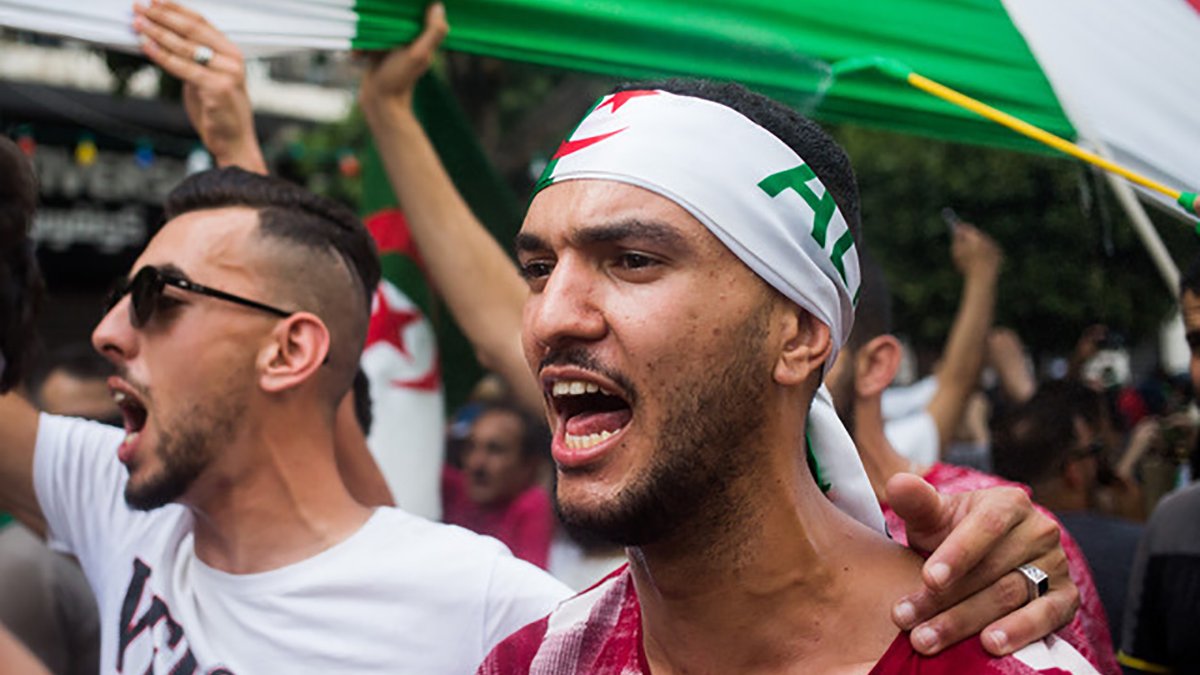 L'image montre des manifestants lors d'une protestation, probablement en lien avec une cause nationale. Deux hommes sont au premier plan : l'un porte un bandana avec des motifs évoquant l'Algérie et semble exprimer de la passion en criant. L'autre homme, à sa gauche, affiche une attitude engagée tout en portant un t-shirt à motifs. En arrière-plan, on peut voir un groupe de personnes brandissant des drapeaux, créant une ambiance de solidarité et de revendication. Les visages sont chargés d'émotion et l'atmosphère paraît énergique.