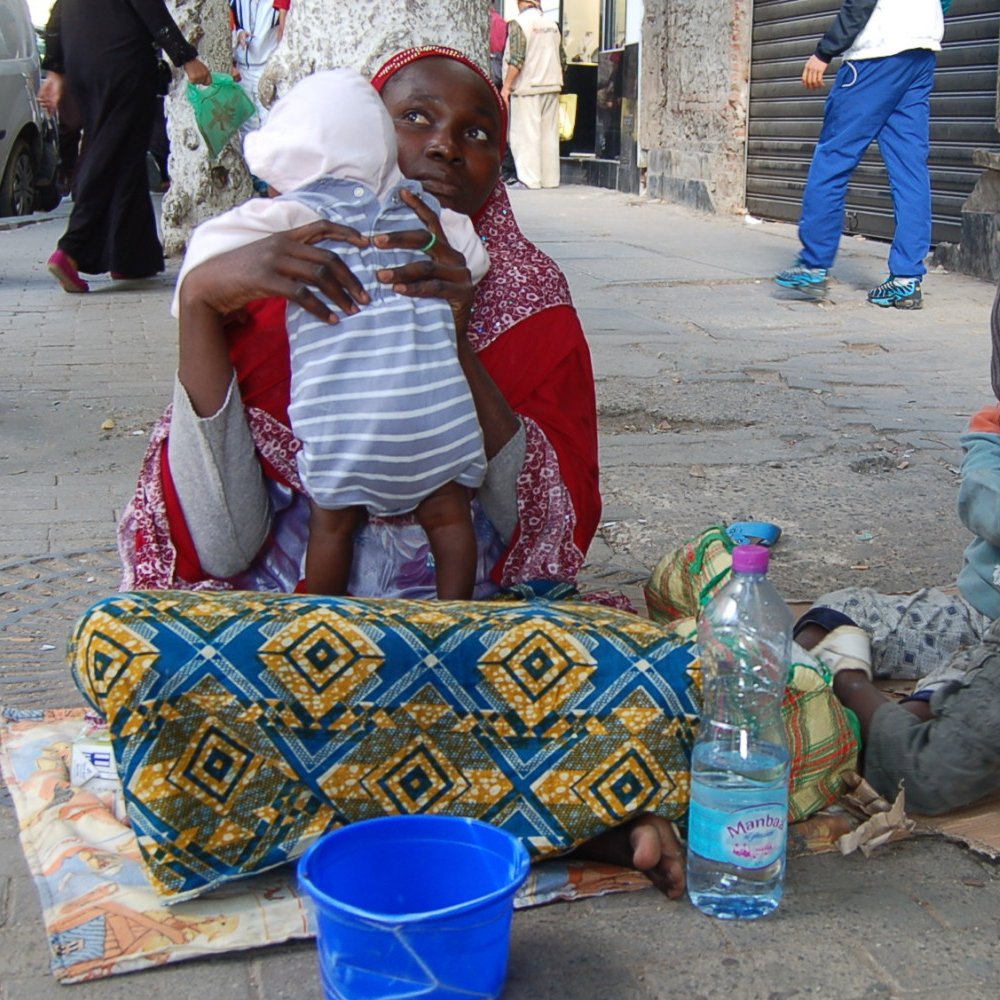 The image depicts a scene on a city street where a woman is seated on the ground with a baby in her arms. She appears to be wearing traditional clothing, characterized by vibrant colors and patterns. Nearby, two young children are playing on the pavement; one is sitting in a casual posture, while the other is occupied with an object. There is a blue bucket beside them and a plastic bottle of water. The background includes other people walking by and buildings that suggest an urban environment.