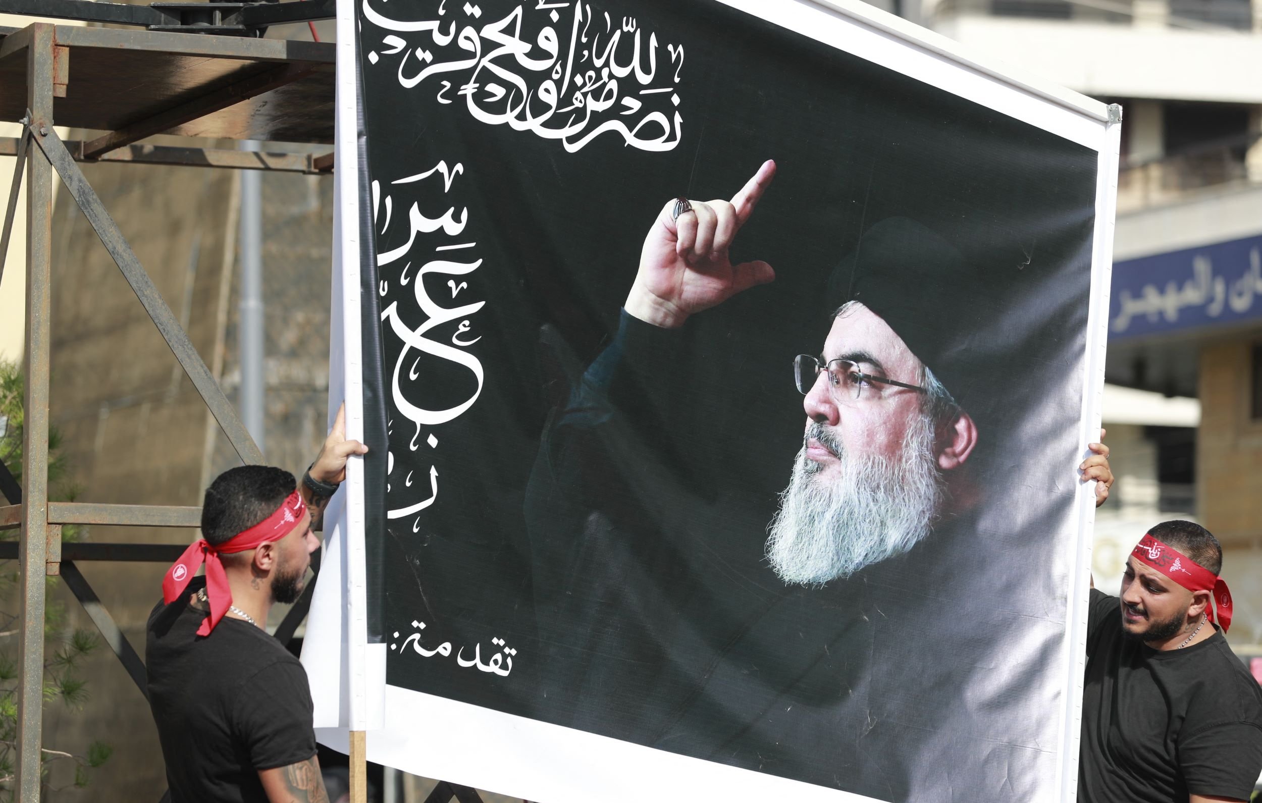 L'immagine mostra due uomini che sorreggono un grande striscione. Sullo striscione è rappresentato il volto di un uomo con un gesto della mano che sembra indicare qualcosa. Il contesto suggerisce una celebrazione o una manifestazione, con i partecipanti che indossano fasce rosse. Lo sfondo è urbanizzato, con edifici visibili.