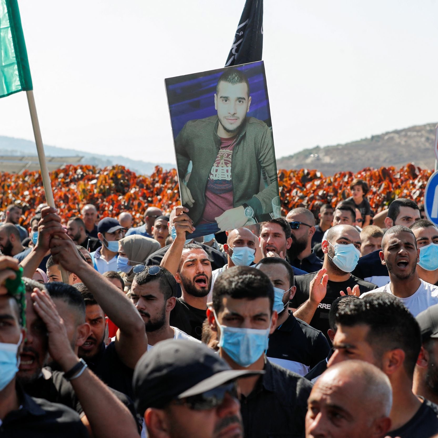 The image depicts a large crowd participating in a protest or demonstration. Many individuals are holding up flags, including green ones, and a prominent figure is holding a portrait of a man. The crowd appears to be engaged and unified, with some people wearing face masks. The background features hills, adding to the overall atmosphere of the event.