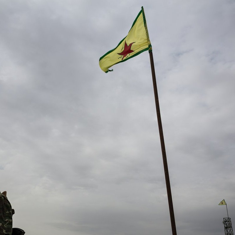 L'image montre un homme en uniforme militaire se tenant près d'un drapeau. Le drapeau est de couleur jaune avec une étoile rouge au centre, entouré de bandes vertes. L'arrière-plan est nuageux, ce qui donne un ton sombre à la scène. En plus de l'homme, on peut apercevoir une autre instance du drapeau en arrière-plan, également hissé sur un mât.