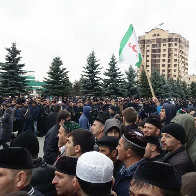 L'image montre une grande foule rassemblée dans une rue, probablement lors d'une manifestation ou d'un rassemblement public. Les participants semblent en majorité masculins et portent différents types de couvre-chefs. Certains tiennent des drapeaux, et l'atmosphère semble mobilisée. En arrière-plan, on peut voir des bâtiments et des arbres, ce qui donne un aperçu de l'environnement urbain. Le ciel est nuageux, ce qui pourrait indiquer un temps frais.