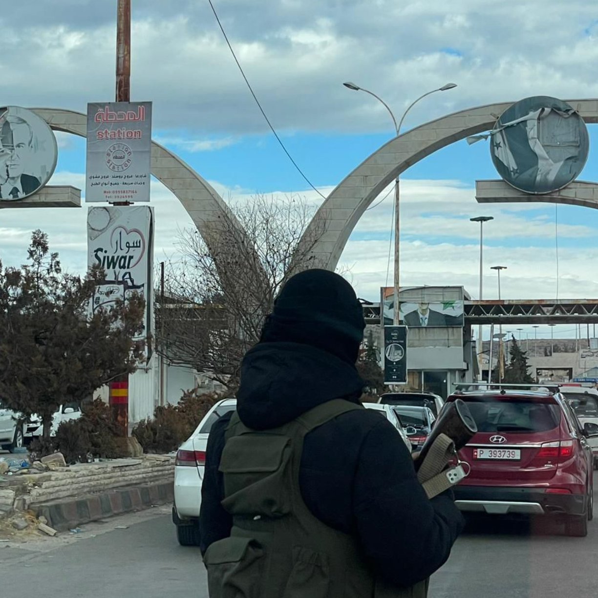 L'image montre un grand arc de triomphe à l'entrée d'une ville, avec des réparations visibles. Deux panneaux ronds sont accrochés en haut, affichant des illustrations. La scène est animée avec des voitures et des passants, sous un ciel nuageux.