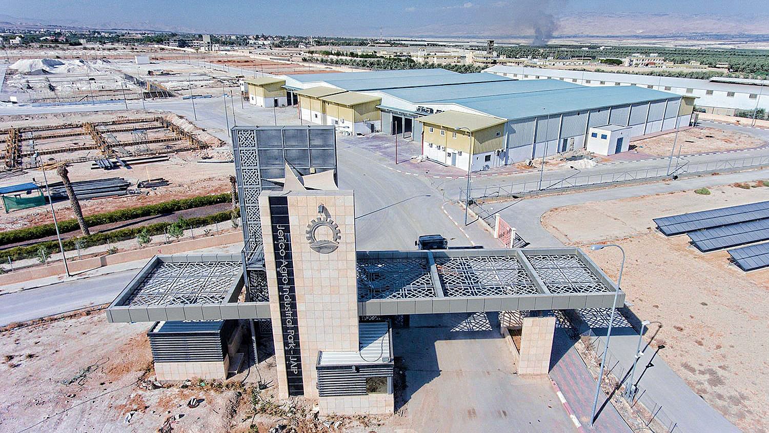 تظهر الصورة مشهداً جوياً لمدخل منشأة أو منطقة صناعية. يتوسط المدخل بناء حديث ذو تصميم معماري مميز، مع واجهة تحتوي على عناصر فنية تتضمن الزخارف والأشكال الهندسية. خلف المدخل، تمتد المستودعات والمبانِ الصناعية، مع تواجد أرضيات مرصوفة ومناطق خضراء ذات أشجار. في الأفق، يمكن رؤية دخان يخرج من أحد المواقع، مما يشير إلى نشاط صناعي، بينما يتواجد في الجانب الأيسر ألواح شمسية. الجو العام يبدو هادئاً ومنظمًا، مع مساحات شاسعة من الأرض.