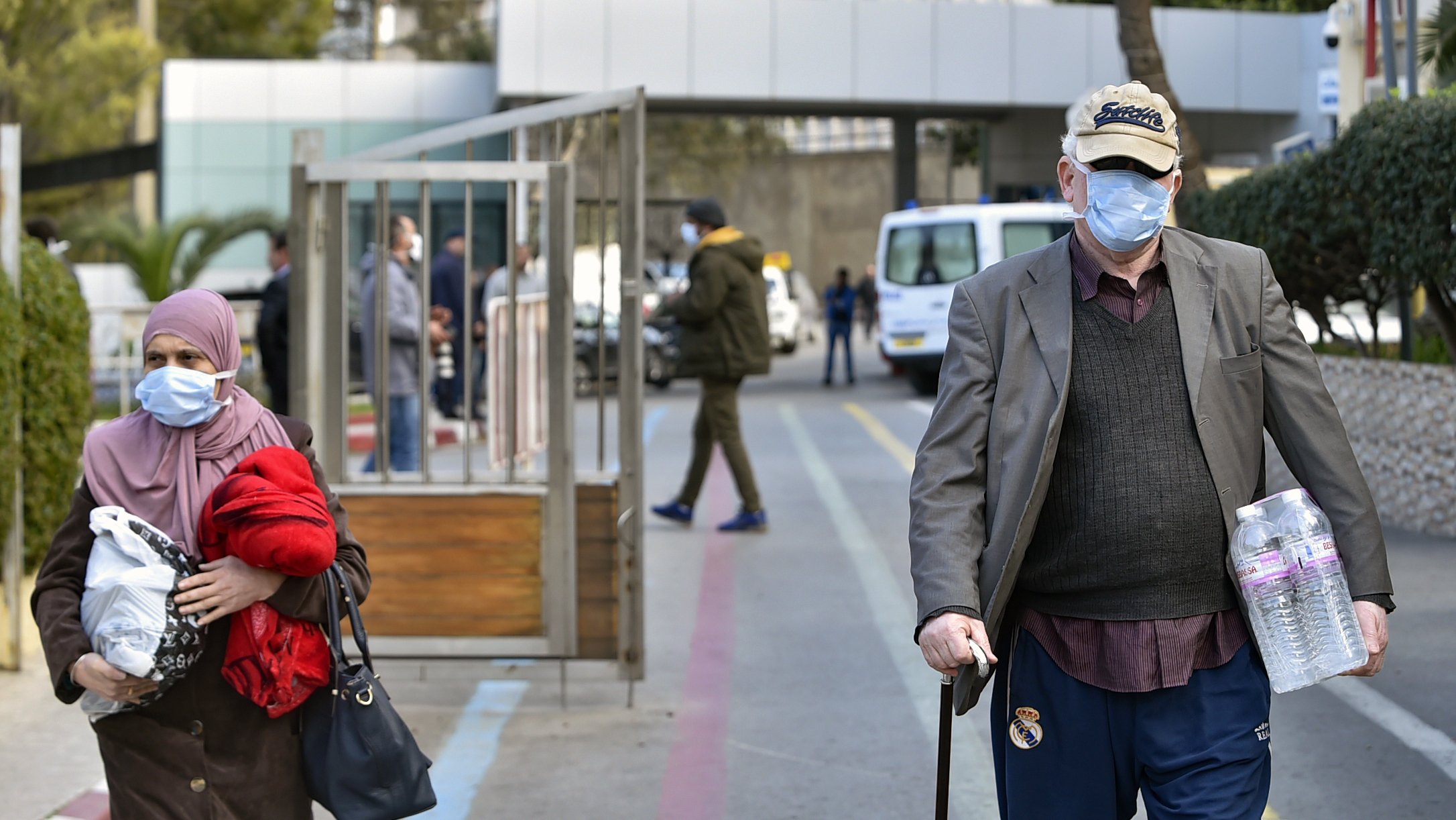 L'image montre une scène urbaine où deux personnes marchent dans une rue. À gauche, une femme portant un foulard et une veste rouge tient un sac, tandis qu'à droite, un homme âgé, avec une canne et un chapeau, marche en portant deux bouteilles d'eau. Les deux portent des masques faciaux, suggérant un contexte sanitaire. En arrière-plan, on aperçoit des véhicules et des bâtiments.