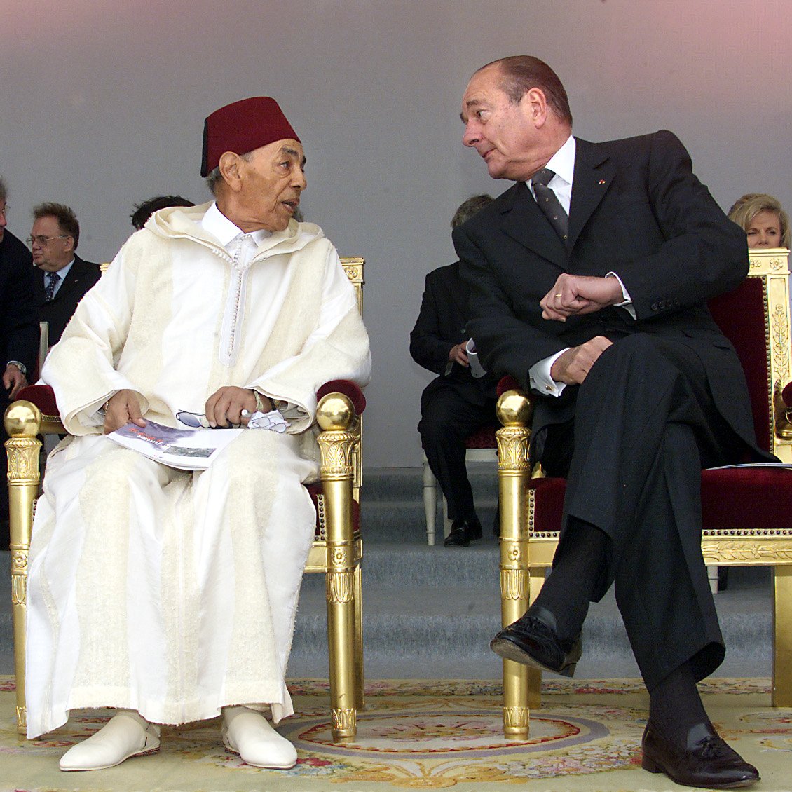 L'image montre deux hommes assis sur des chaises ornées, dans un cadre formel. L'un porte un habit traditionnel marocain avec un fez rouge, tandis que l'autre est vêtu d'un costume noir. Ils semblent engagés dans une conversation, se regardant avec attention. En arrière-plan, on aperçoit d'autres personnes, indiquant que cette scène a lieu lors d'un événement officiel. Leurs expressions semblent amicales et concentrées.