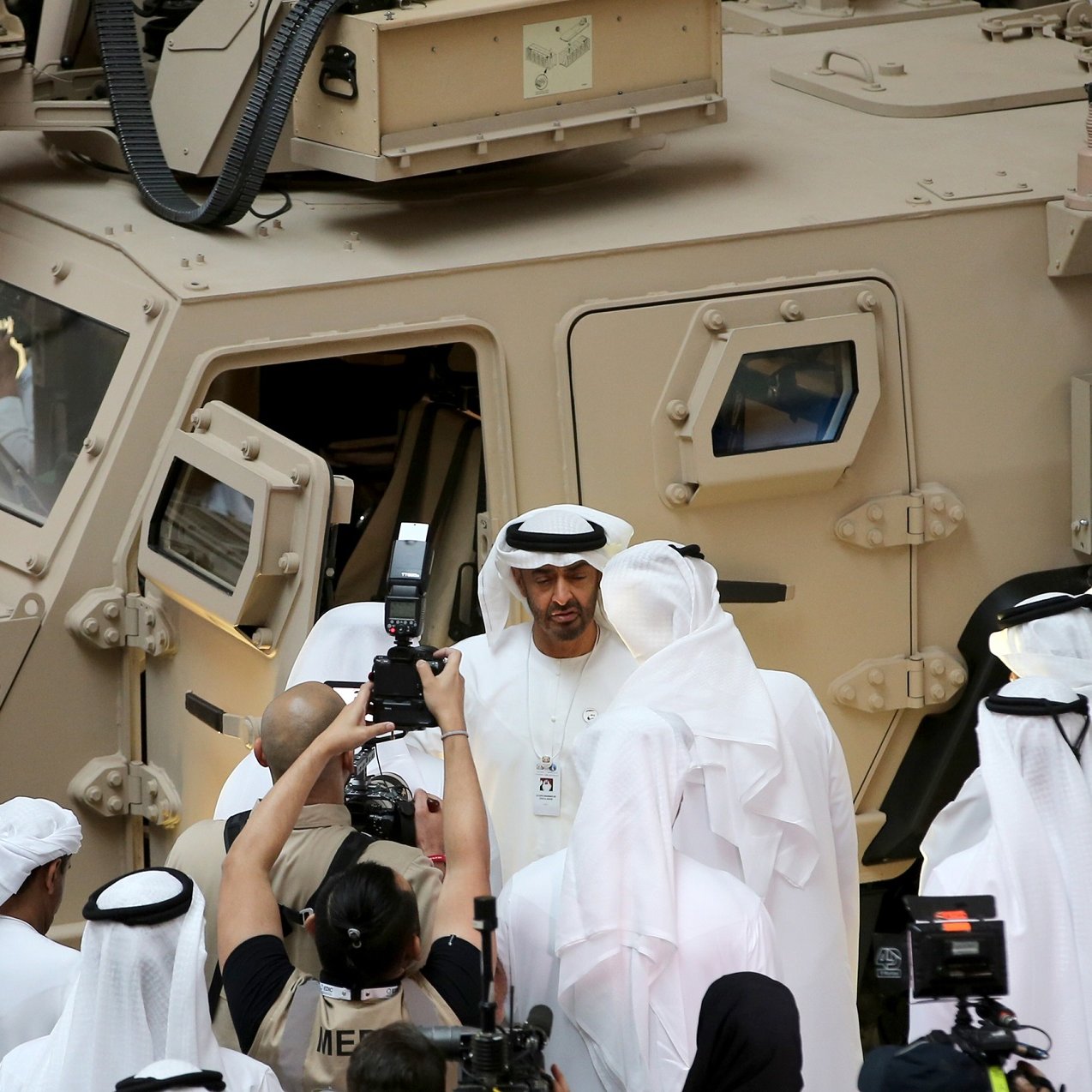 L'image montre un groupe de personnes, probablement des dignitaires, réunis autour d'un véhicule militaire dans un environnement formel. Plusieurs hommes portent des vêtements traditionnels, comme des kandouras et des ghutras. Il y a également des journalistes et des caméras en train de filmer ou de prendre des photos, ce qui suggère un événement médiatique ou une visite officielle. Le véhicule militaire est de couleur beige et semble robuste, avec plusieurs fenêtres et des équipements visibles.